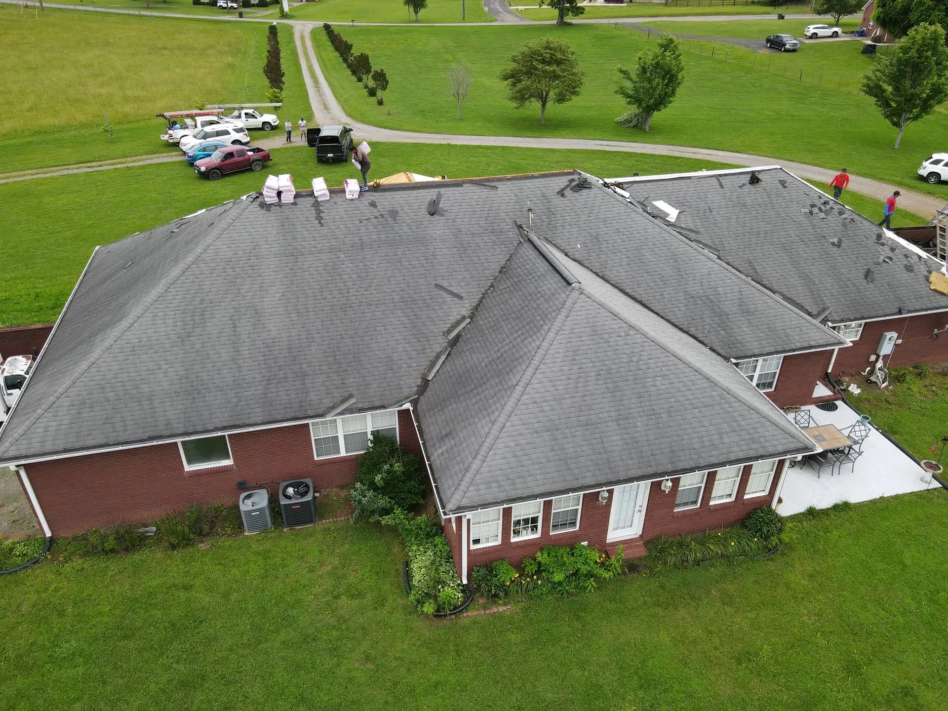Roofers working on a dark gray shingled roof of a red-brown house, surrounded by green grass and trees.