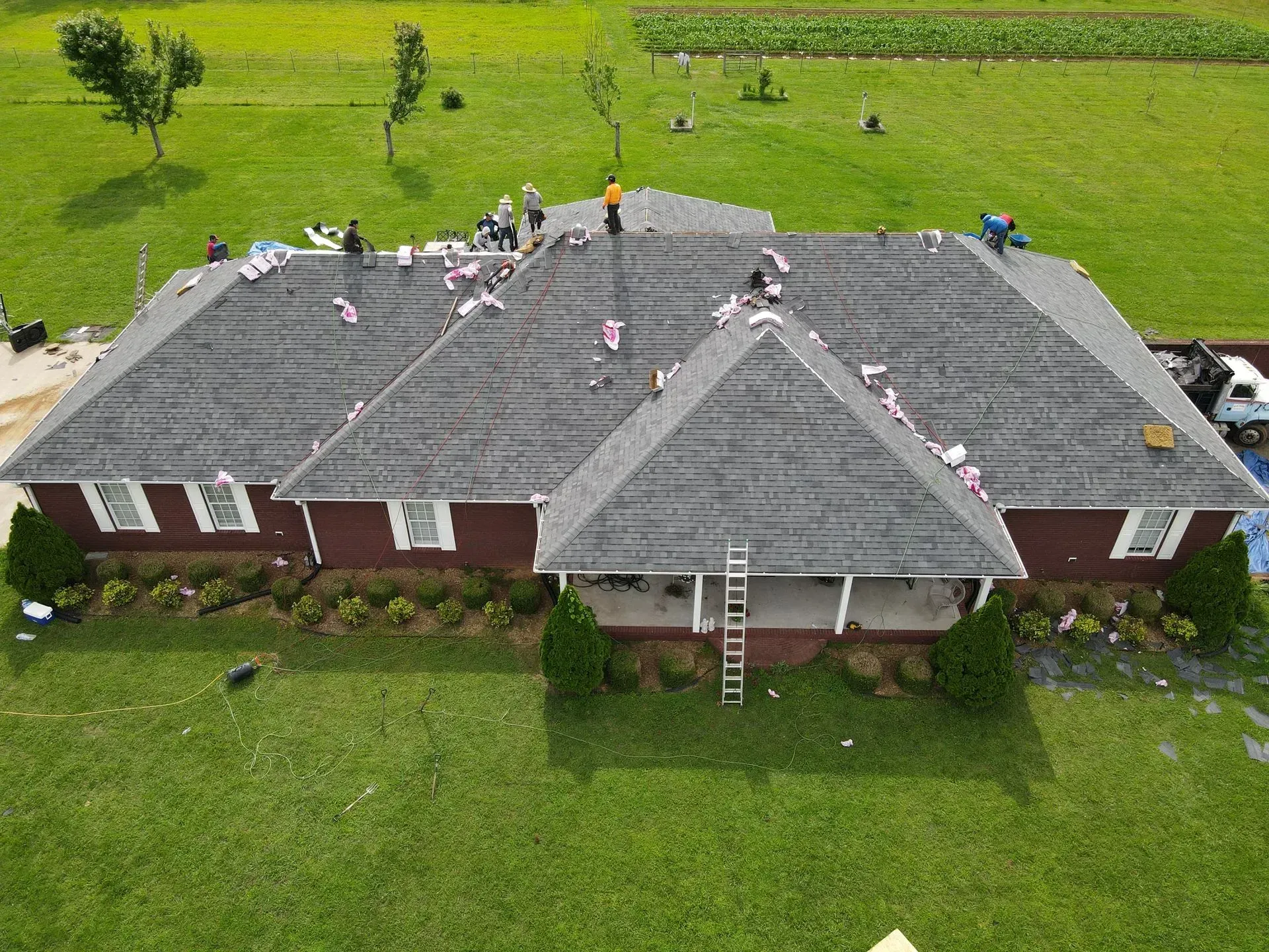Roofing crew working on a dark gray shingled roof of a red brick house in a grassy field.