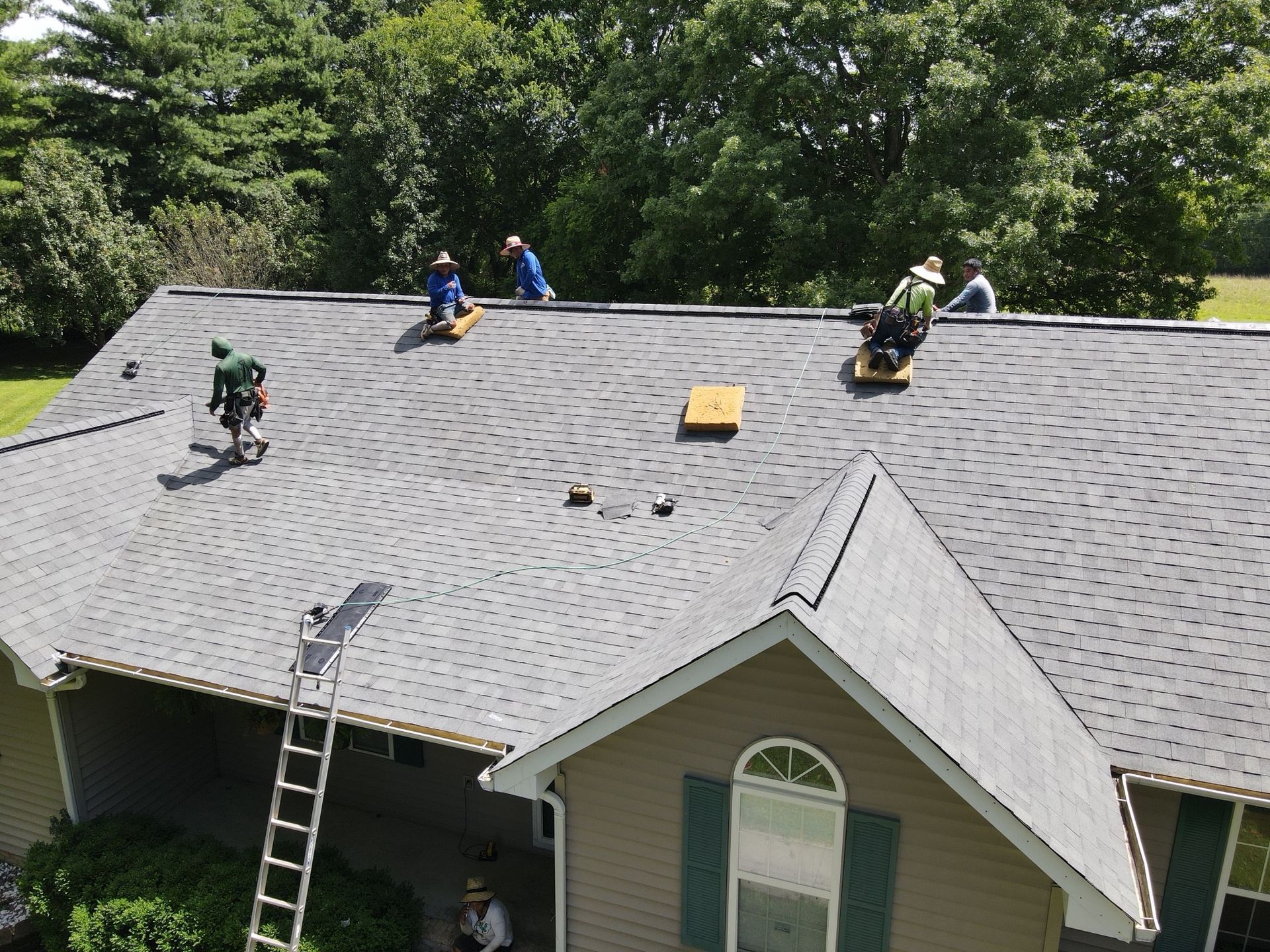 Several workers in hard hats install gray shingles on a suburban house roof under a clear sky.