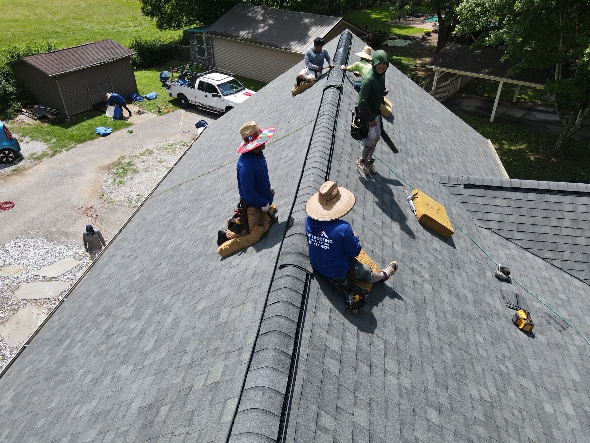 Construction workers in hats and blue shirts install gray shingles on a residential roof.