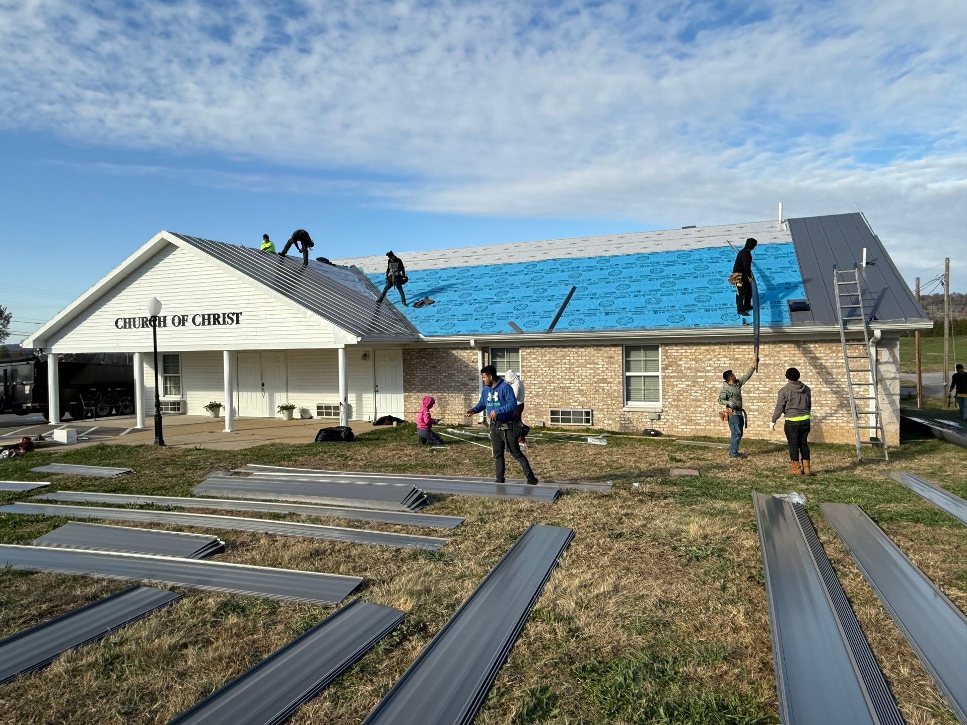Workers replace the roof of a stone-walled church building with metal panels on a sunny day.