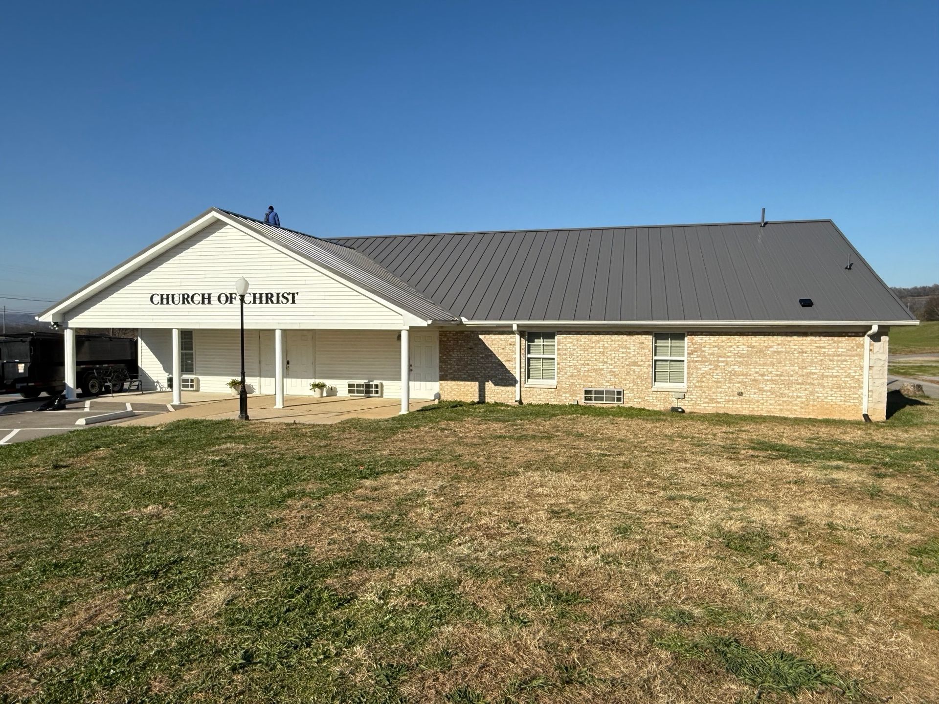 A beige brick building with a dark metal roof and a white covered porch under a clear blue sky.