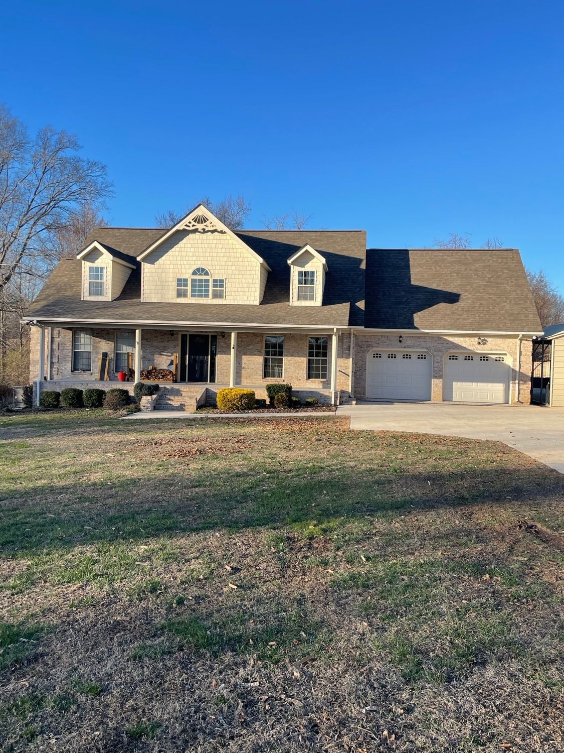 A two-story tan brick house with a dark shingle roof, a front porch, and an attached two-car garage under a blue sky.