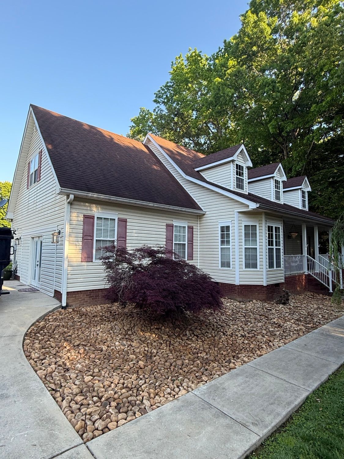 A two-story, white-sided house with a brown shingled roof, three dormers, and a rock-covered front yard under a blue sky.