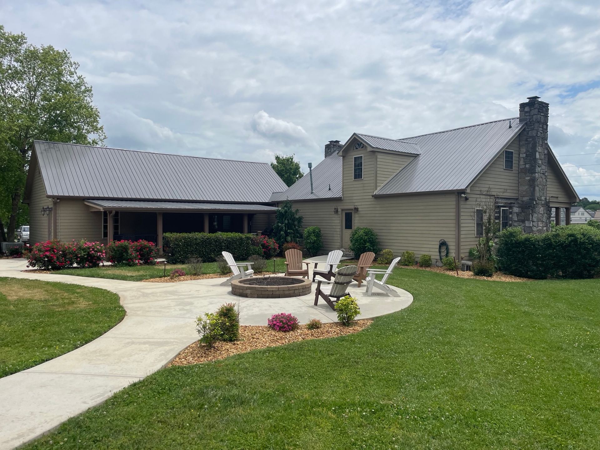 A light brown building with a metal roof, featuring a circular stone fire pit and chairs on a patio in a grassy yard.