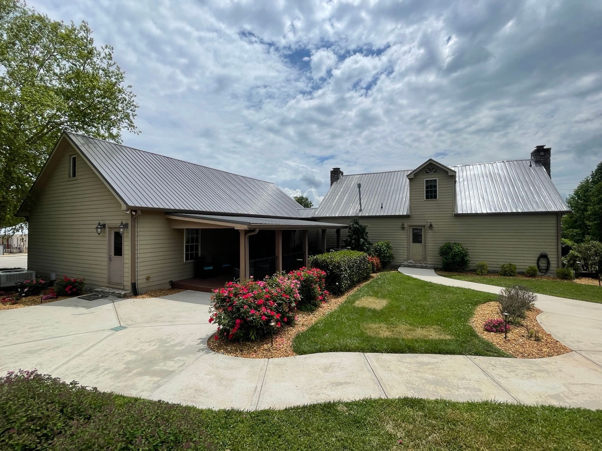 A tan, two-story house with a metal roof, a porch, and a curved concrete driveway surrounded by green grass and bushes.