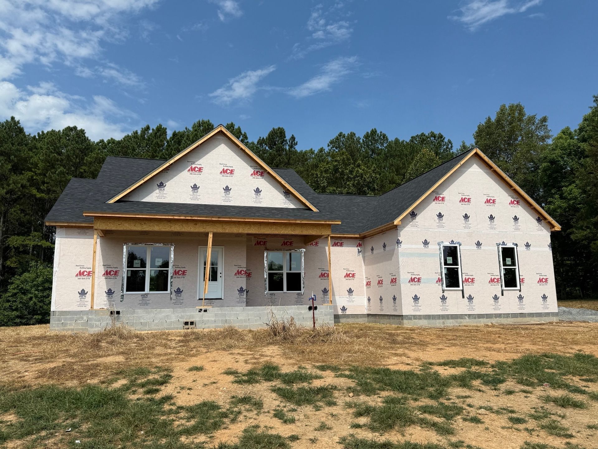 A new house under construction with white sheathing, dark shingles, and installed windows, set against a wooded backdrop.