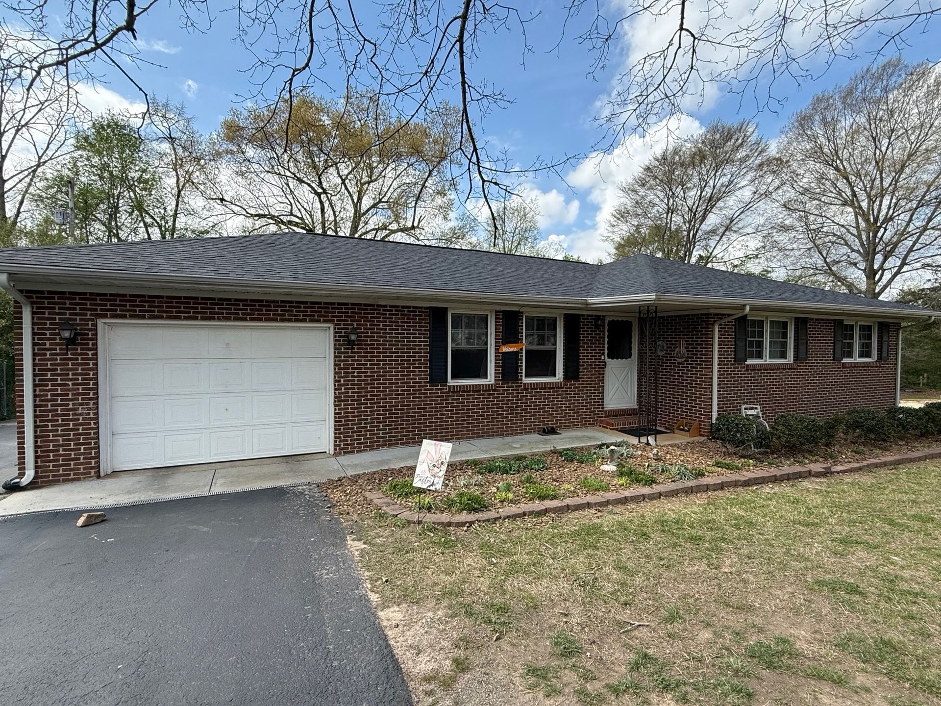 Single-story, dark brick ranch-style house with a gray shingled roof, white garage door, and bare trees under a blue sky.