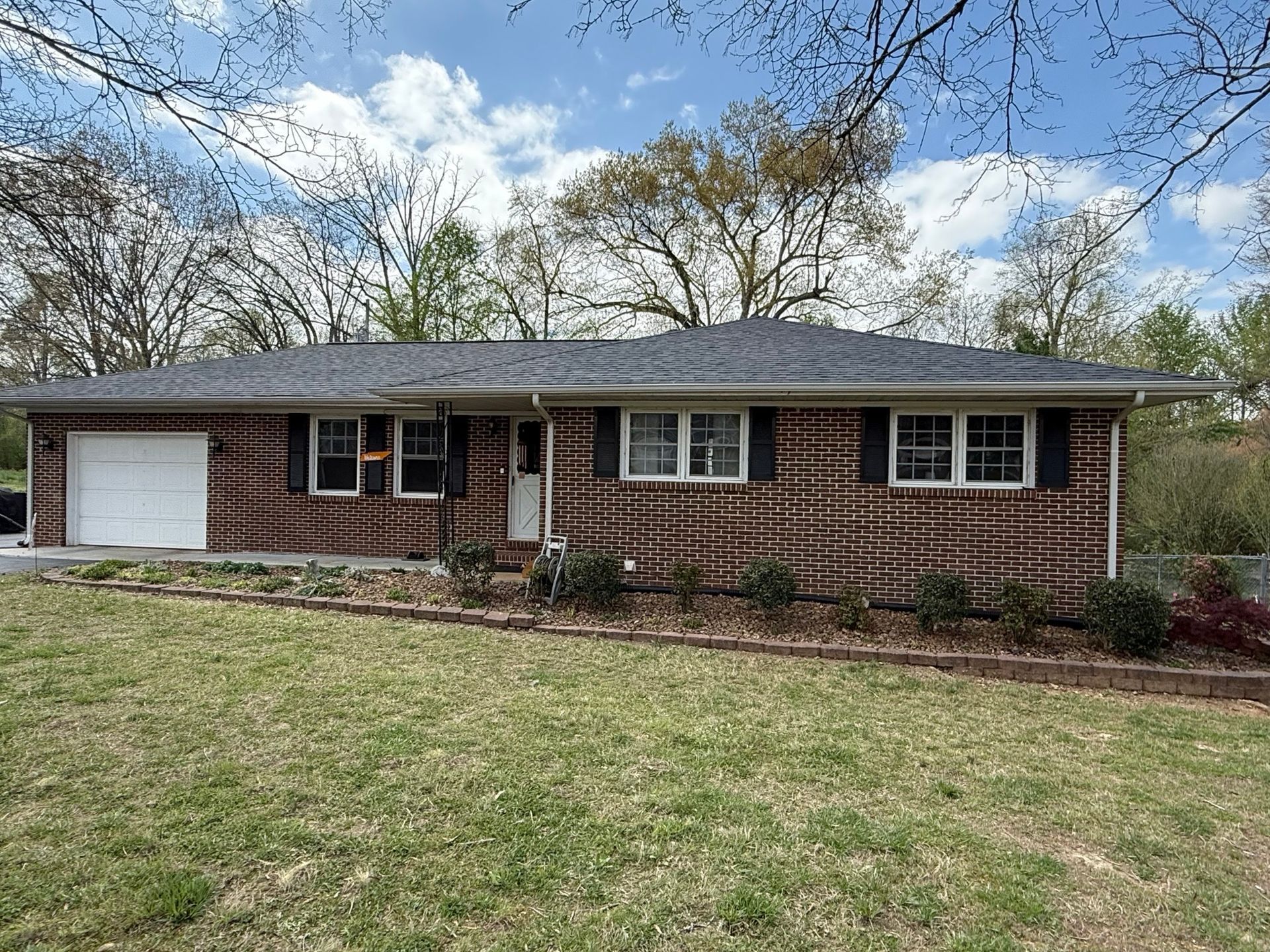 A single-story, brown brick ranch-style house with a white garage door and dark roof, set on a grassy lawn under trees.