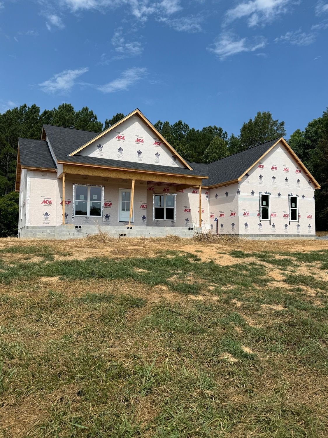 A new single-story house under construction with white sheathing, dark shingles, and a front porch, set in a grassy lot.