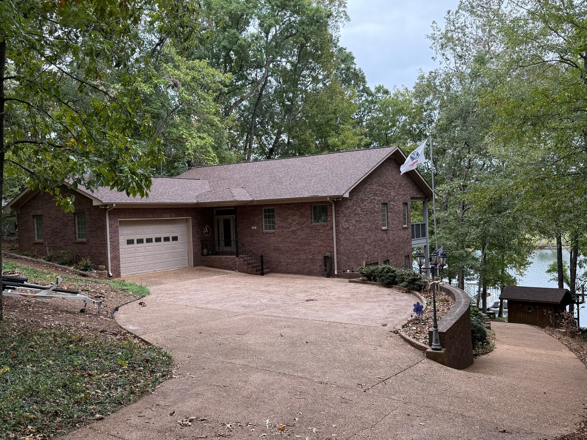 A single-story brick house with a gravel driveway, nestled in a wooded setting overlooking a lake.