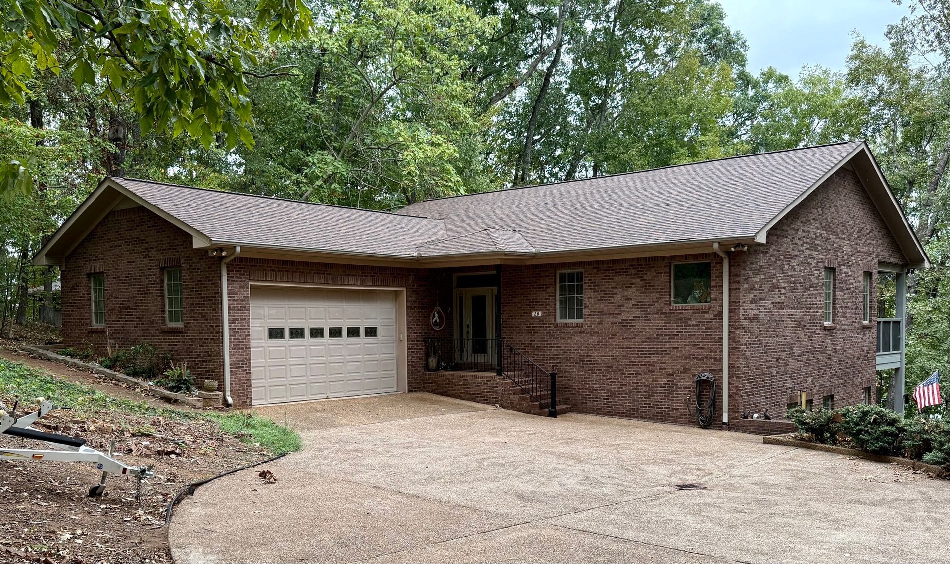 A brown brick, single-story ranch house with a gravel driveway, garage, and surrounding mature trees.