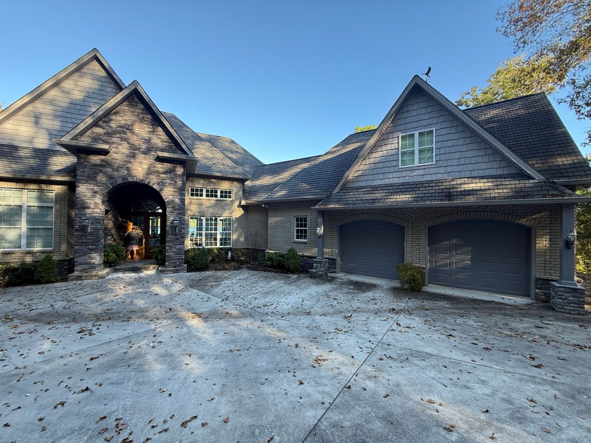 A two-story stone house with a slate roof, arched entryway, and attached two-car garage under a clear blue sky.