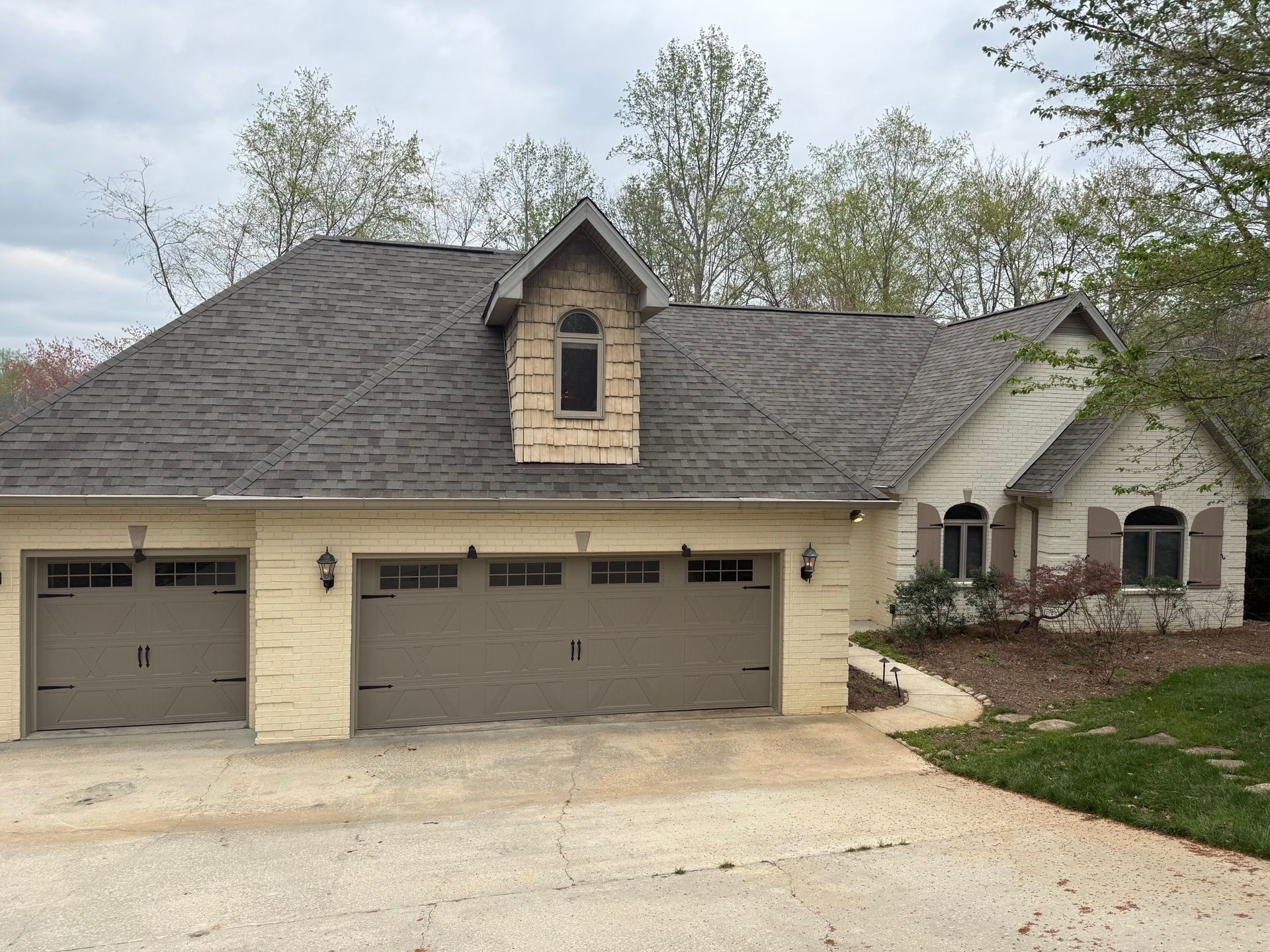 A light-colored stone, three-car garage home with brown shutters, a dormer window, and a gray roof on a paved driveway.