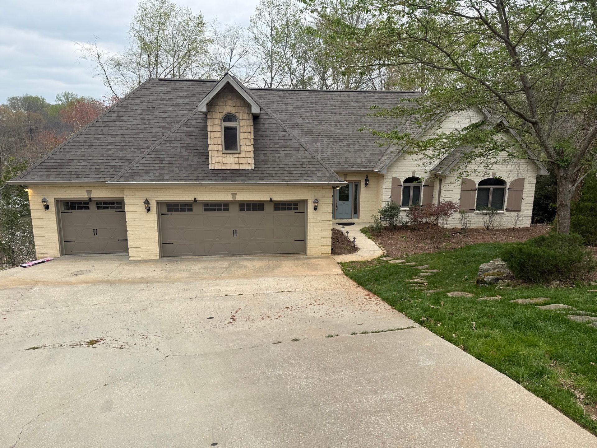 A single-story, beige stone house with a multi-car garage, a dormer window, and a concrete driveway on a grassy lot.