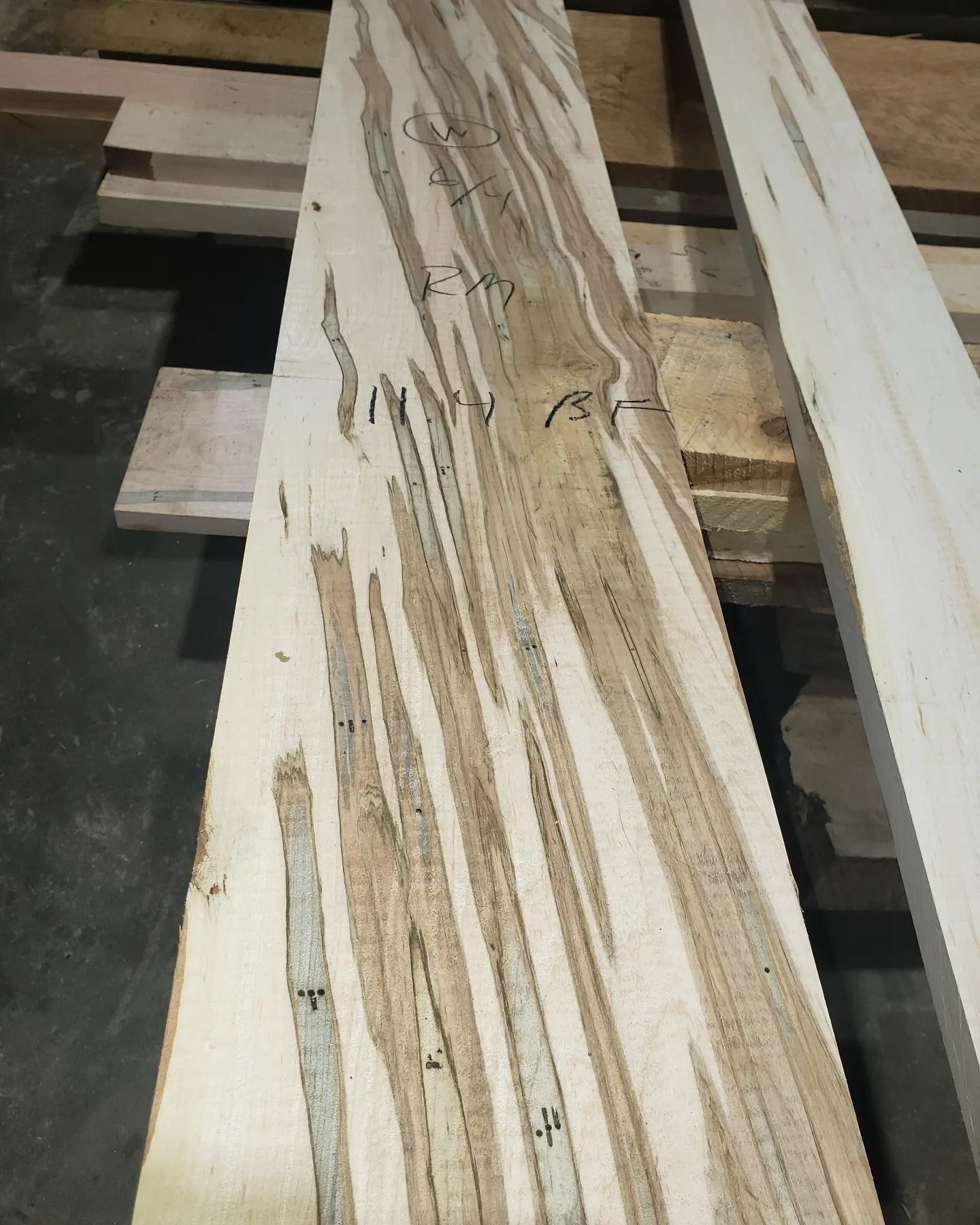 A long, raw wooden plank featuring distinct, dark, mineral-stained streaking patterns across a light cream-colored surface.