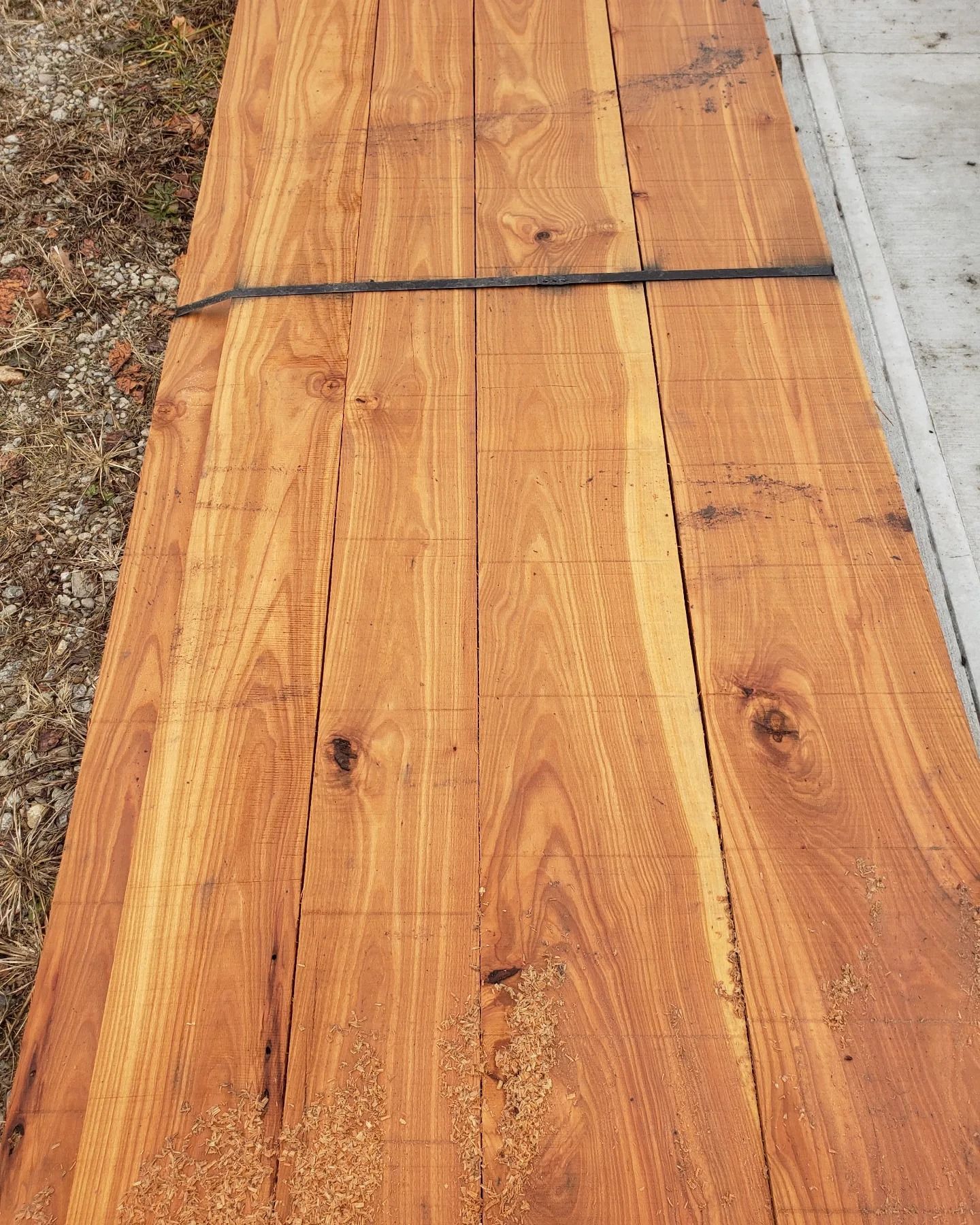 A bundle of rough-cut, light-brown wooden planks lying on gravel next to a concrete edge.