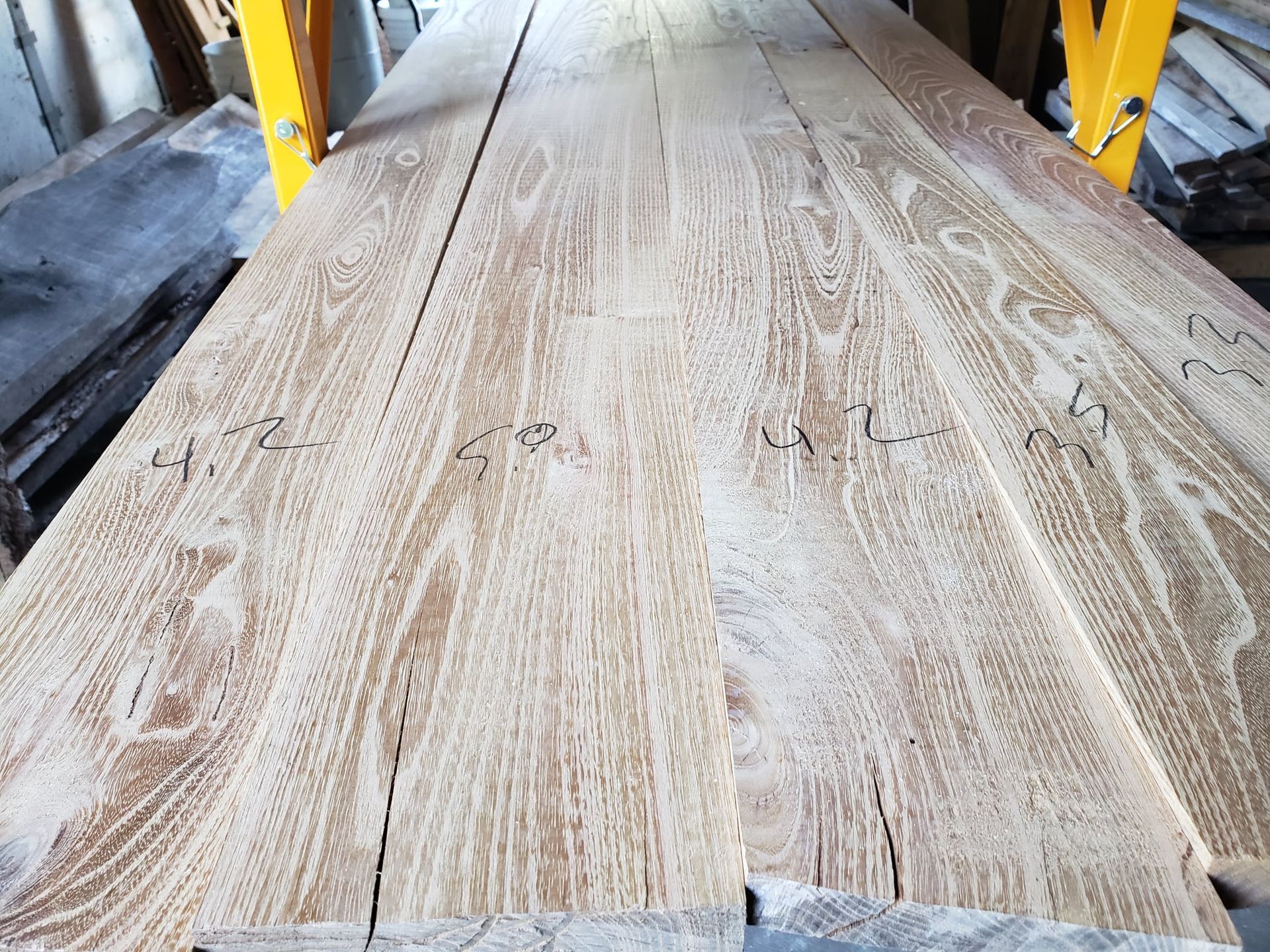 Rough-cut light-colored wooden planks with grain patterns and handwritten numbers resting on a yellow metal rack.