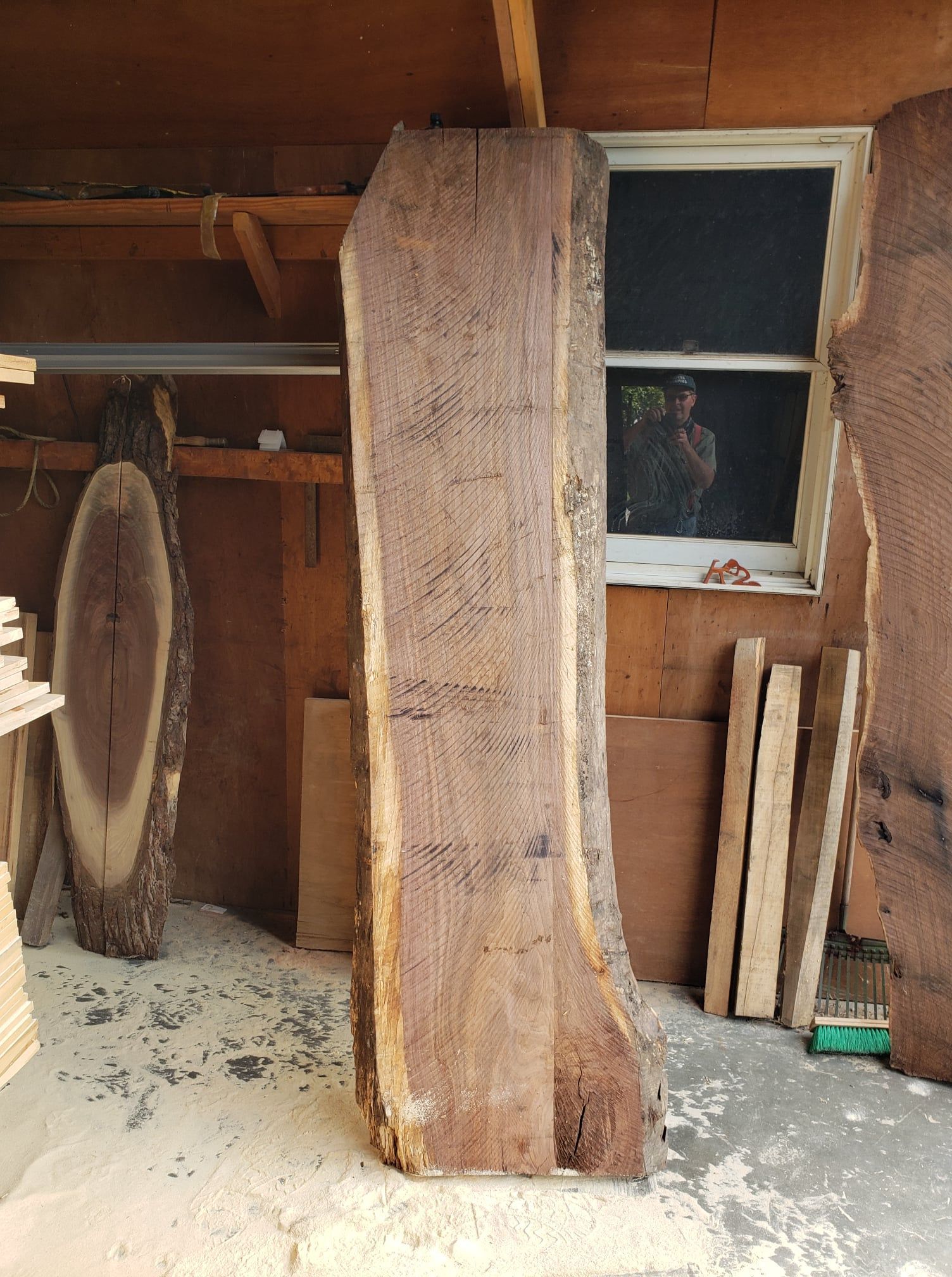 A tall, live-edge wood slab stands vertically in a woodworking shop, with another slab to the left and lumber nearby.
