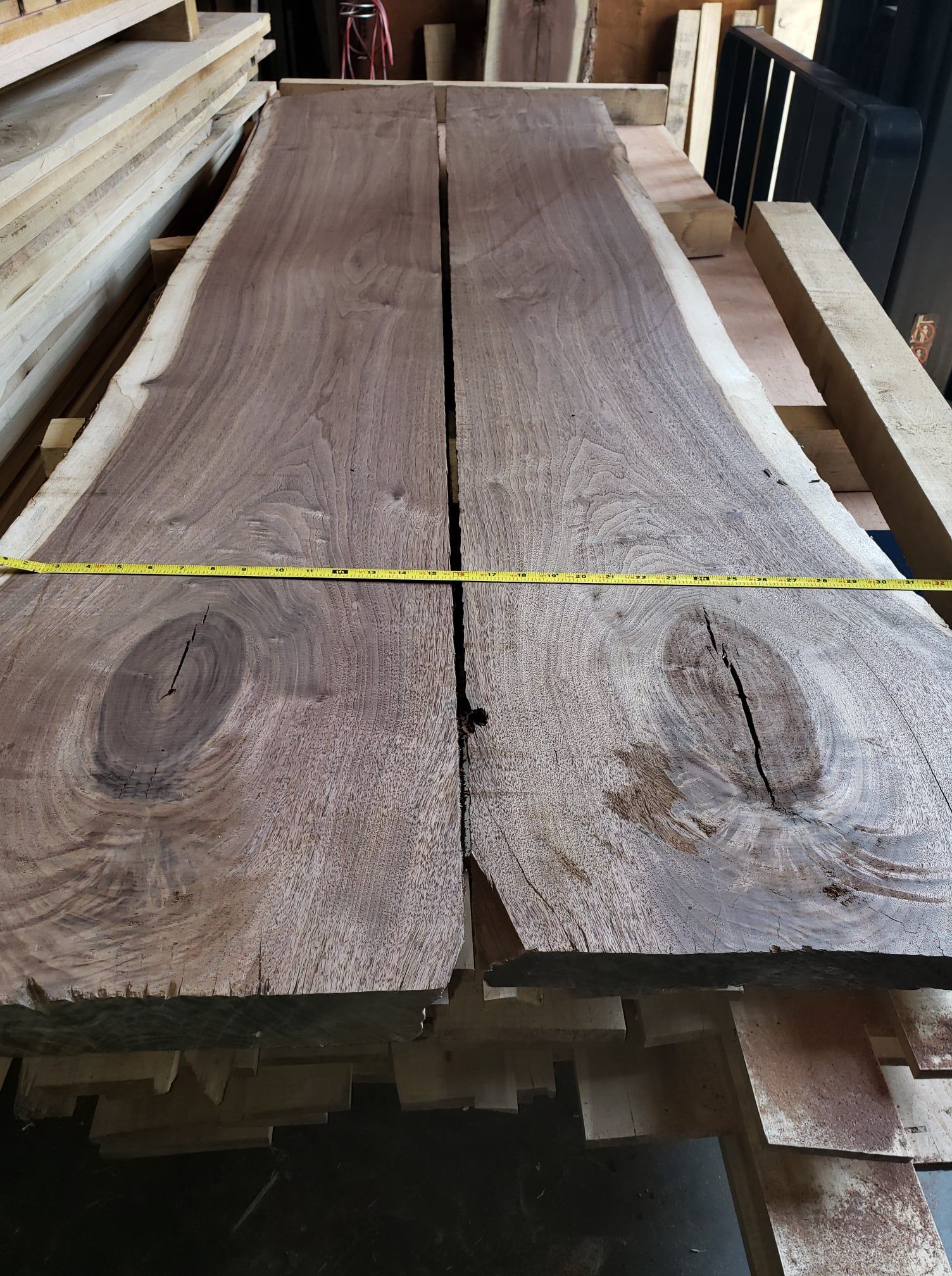 Two rough-cut live edge walnut wood slabs resting on a stack with a yellow measuring tape across them in a workshop.