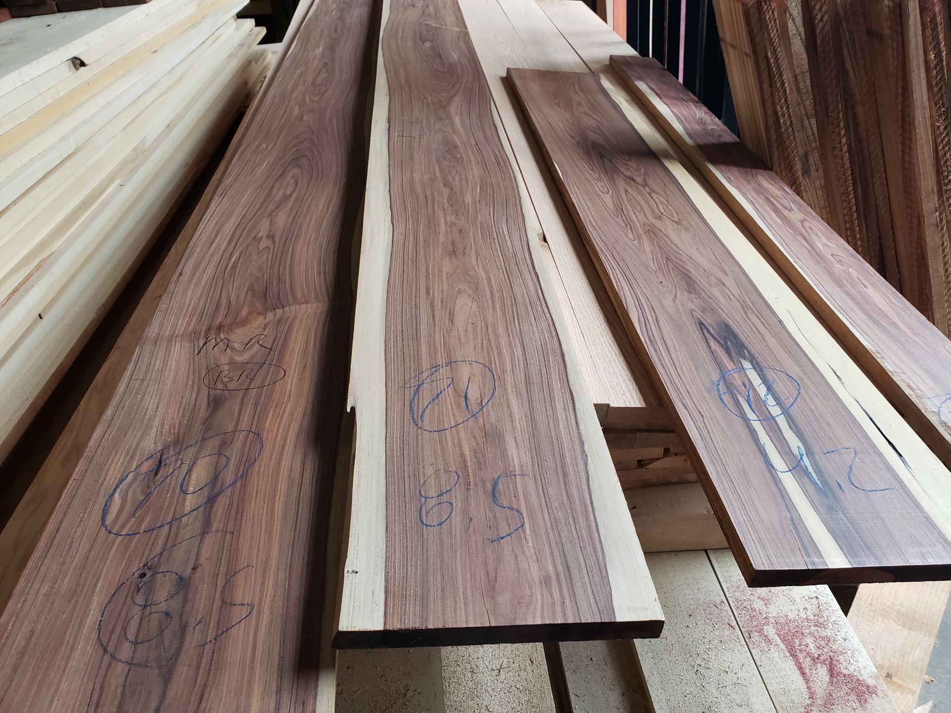 A stack of long, dark brown walnut wood planks with lighter sapwood edges, resting in a lumber yard or workshop.