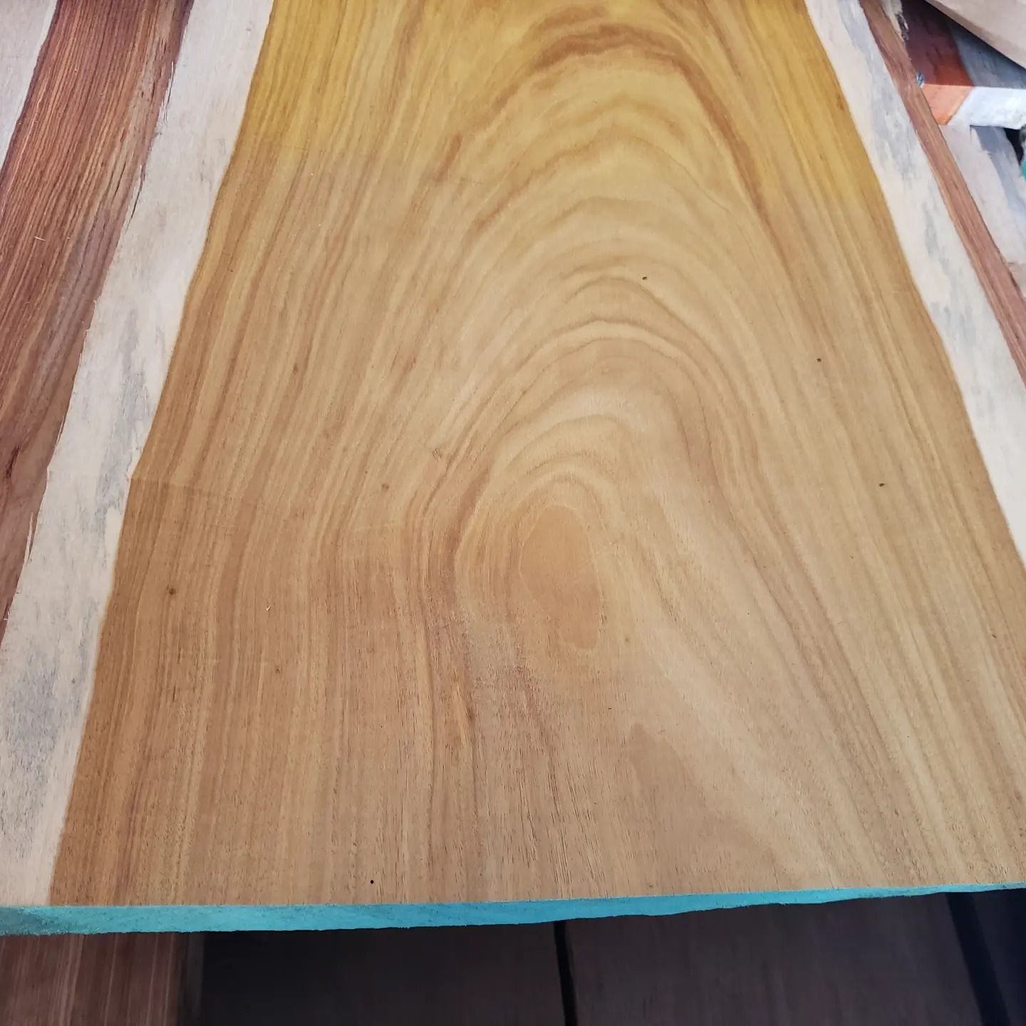 A close-up of a raw wooden board with a light tan center, prominent grain patterns, and lighter-colored sapwood edges.