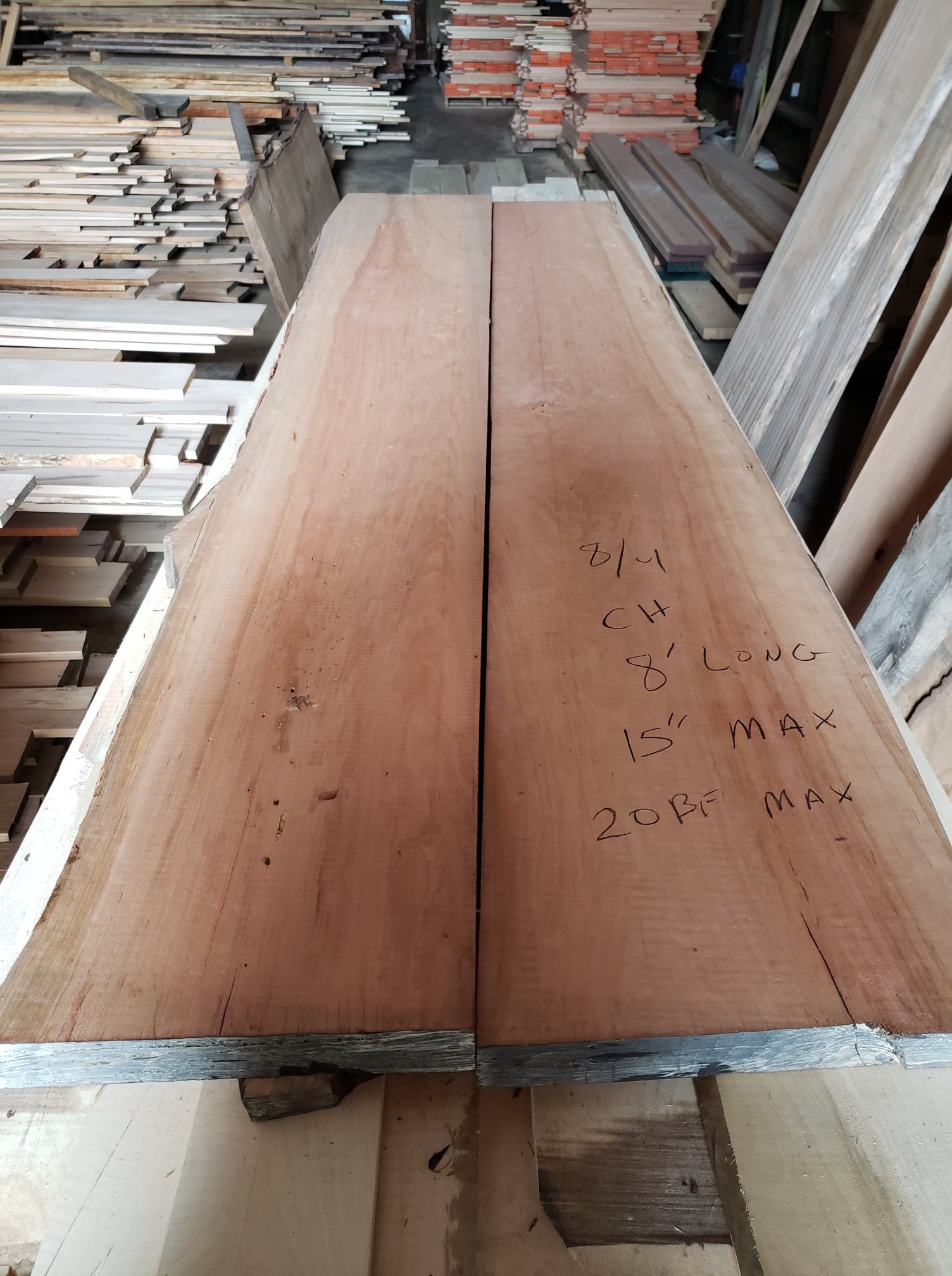 Two long, reddish-brown wooden planks with handwritten specifications sit on a stack of lumber in a workshop.