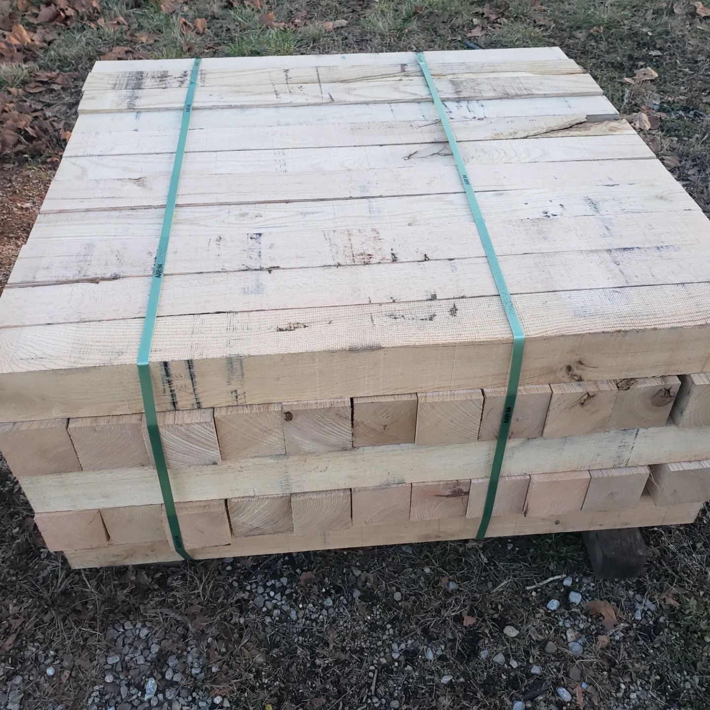 A large stack of light-colored, rough-cut lumber strapped with two green plastic bands sitting on gravelly ground.