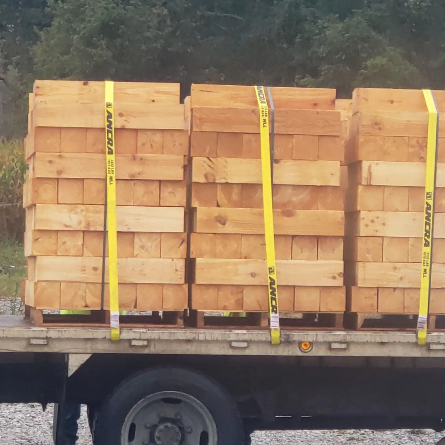 Flatbed trailer loaded with three pallets of stacked lumber, secured by yellow tie-down straps.