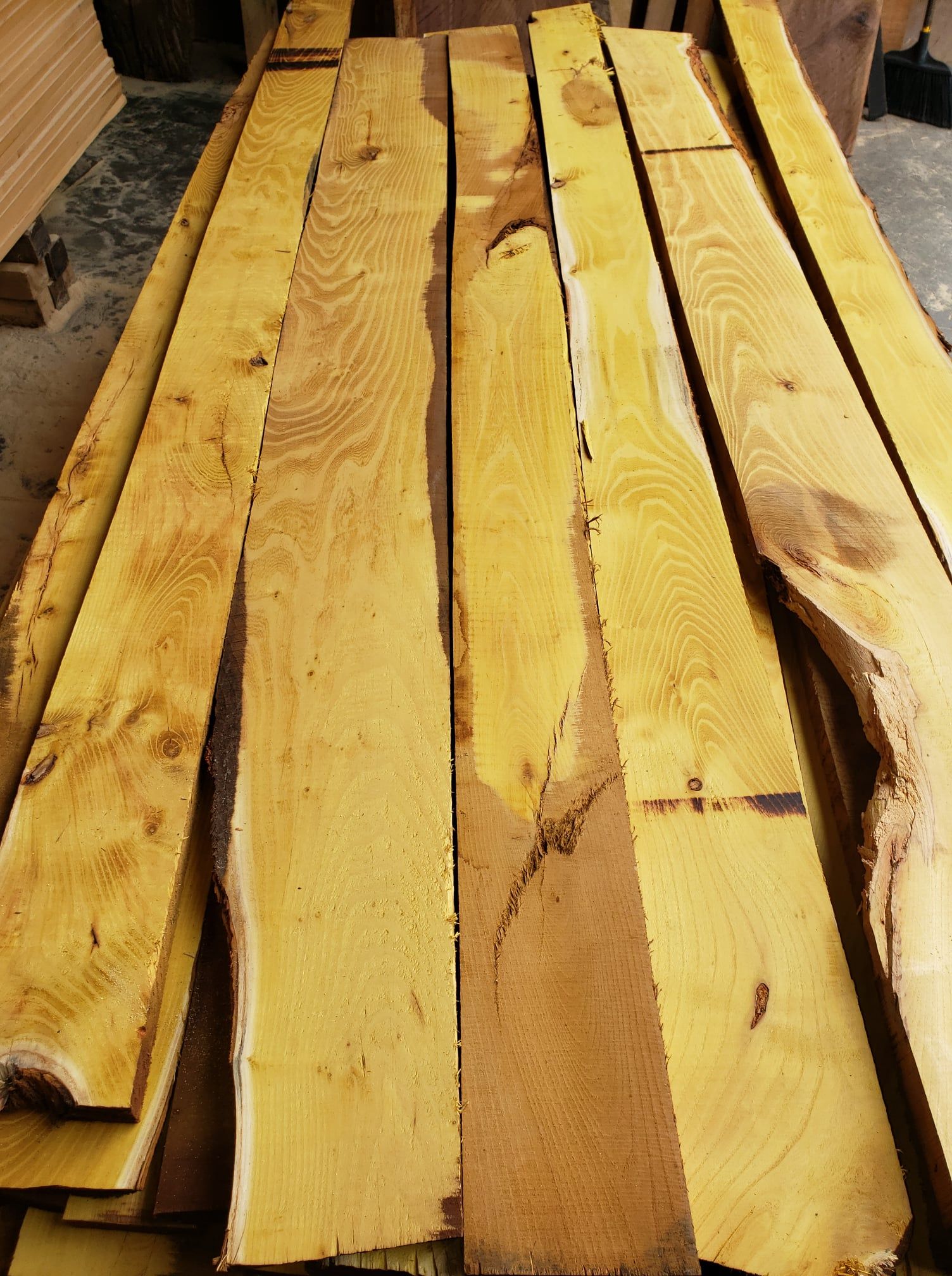 A stack of unfinished, light-yellow wooden boards with rough edges, showing natural wood grain and knots in a workshop.