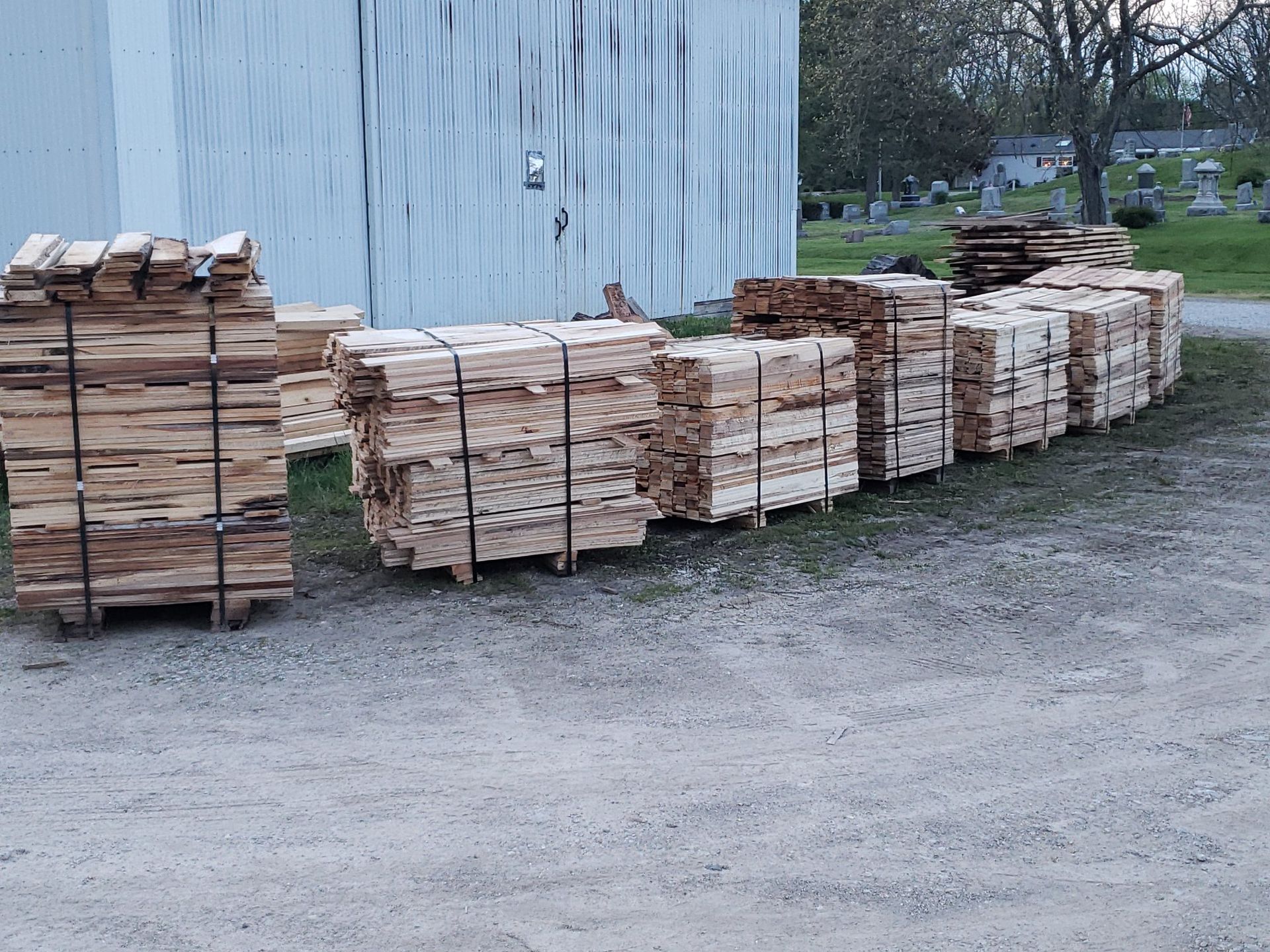 Several stacks of banded lumber are arranged in rows on a gravel lot in front of a white building.