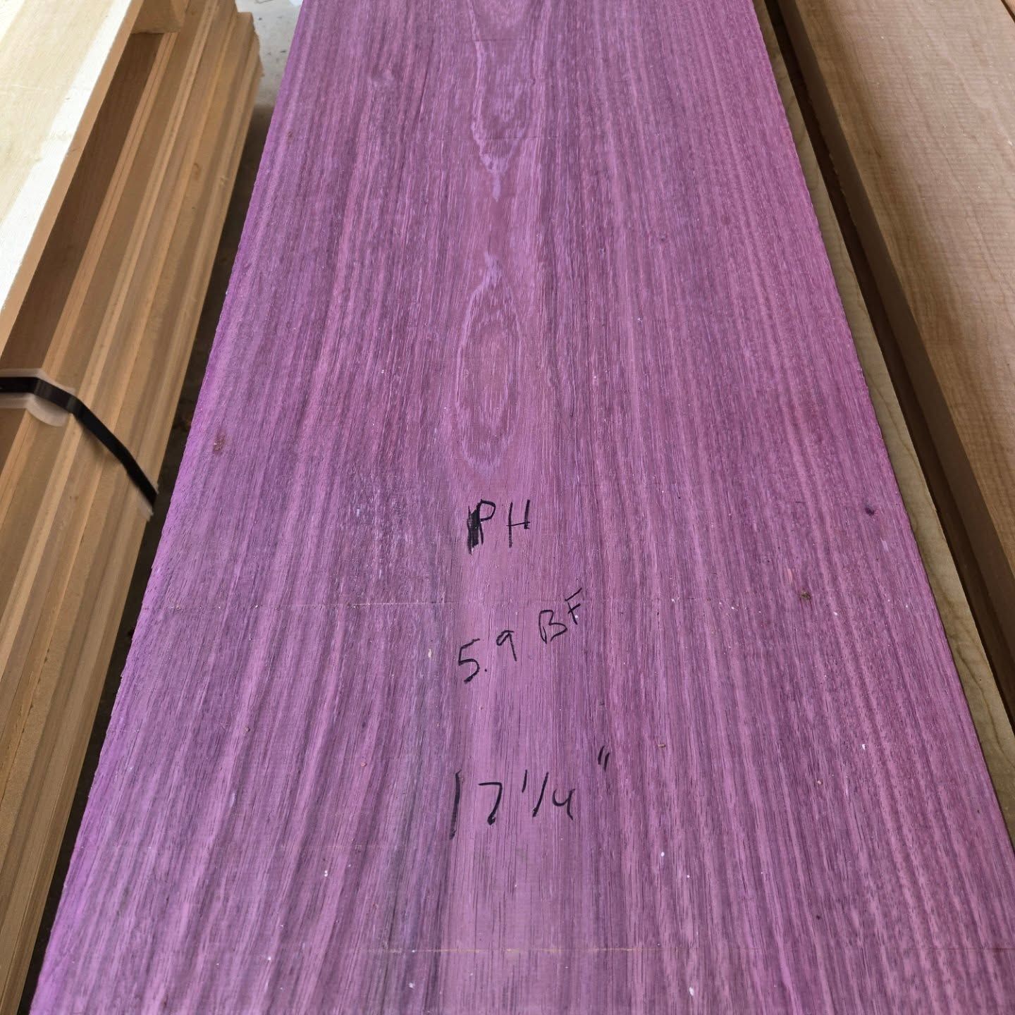 A long board of vibrant purple-colored wood resting among other lumber, with handwritten specifications on the surface.