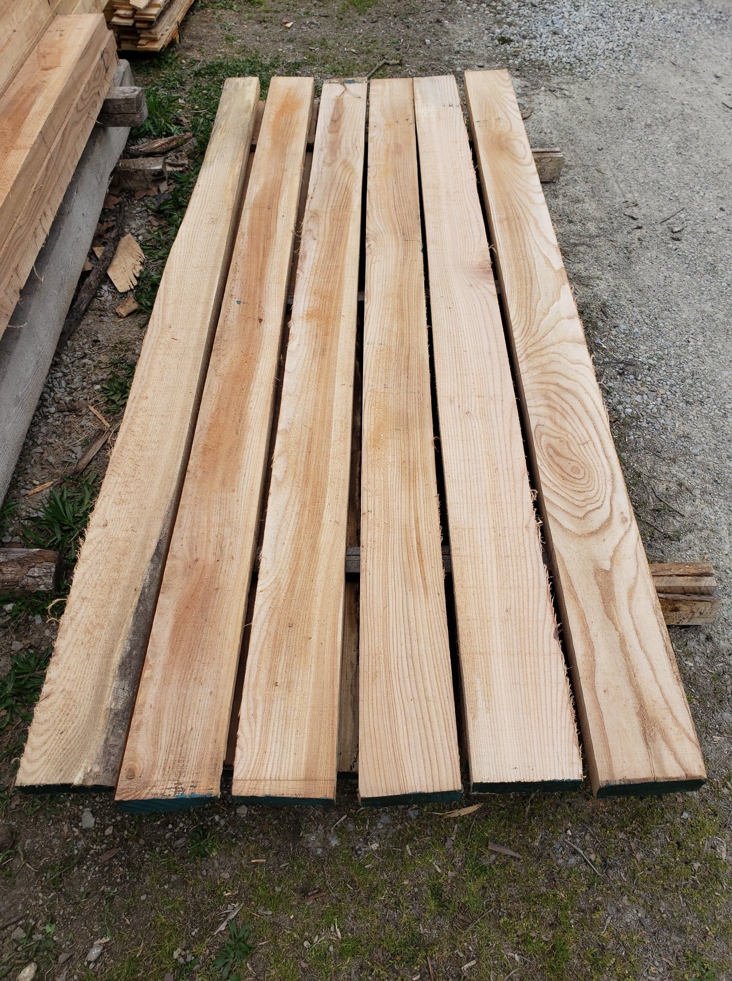 A stack of six long, light-colored wooden planks laid horizontally on a gravel surface outdoors.