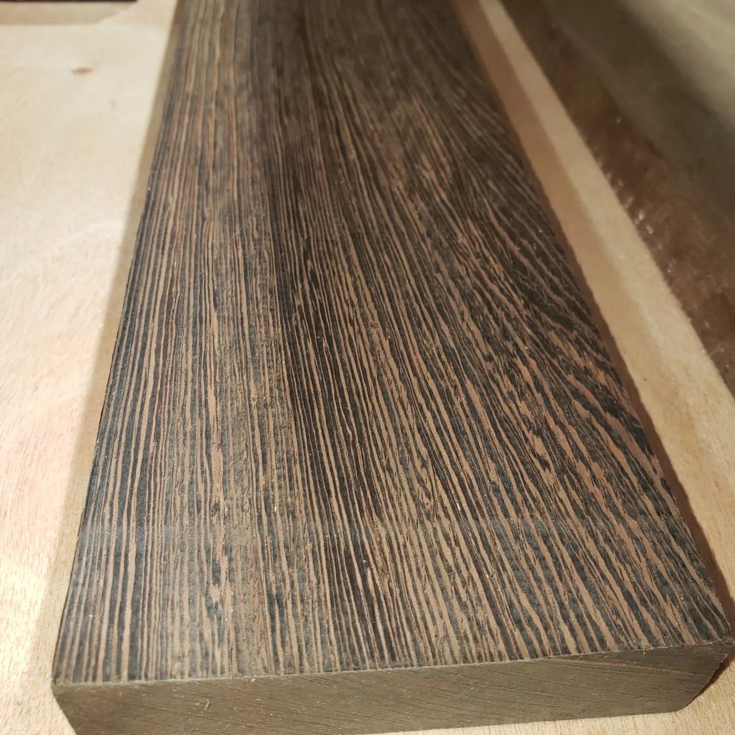 A rectangular plank of dark, textured wood with prominent vertical graining, resting on a lighter wood surface.