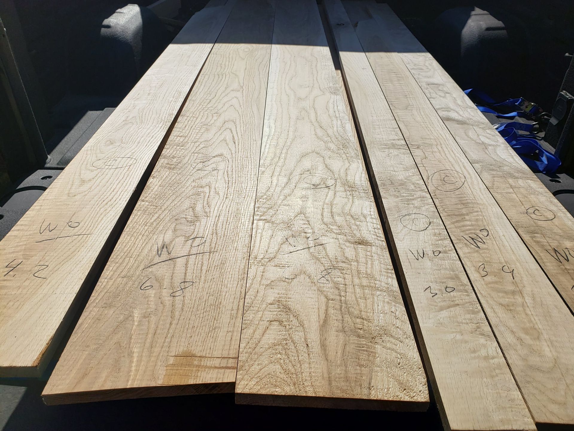 Several long, light-colored wooden planks with handwritten markings lie side-by-side in the bed of a truck.