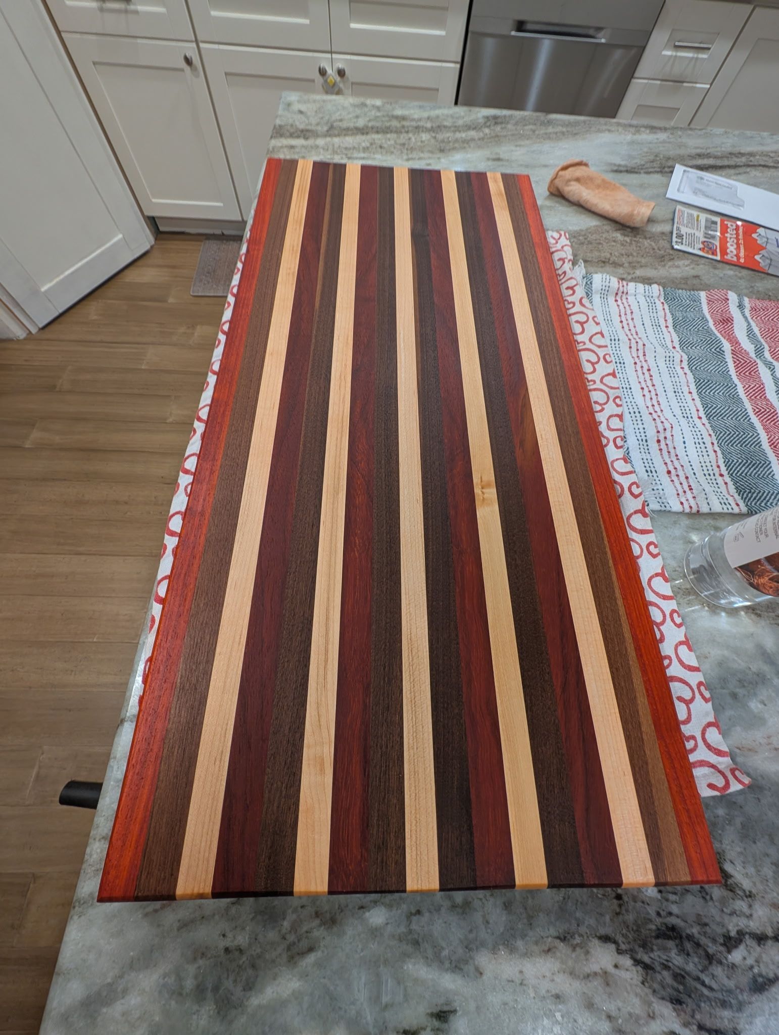 A long, rectangular cutting board with striped patterns in shades of brown, tan, and red sits on a stone kitchen counter.