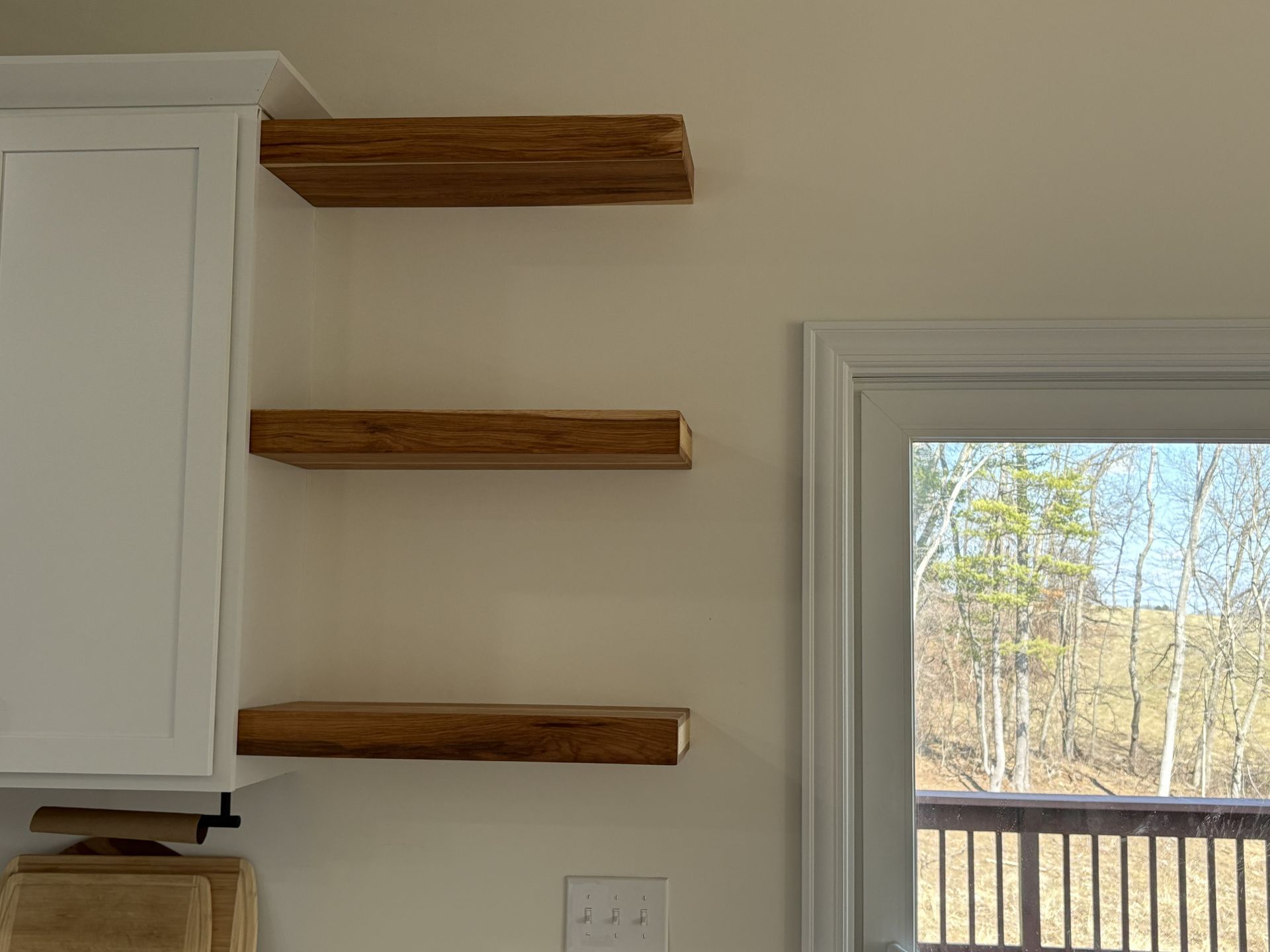 Three wooden floating shelves mounted on a light-colored wall next to a white kitchen cabinet and a door with a view.