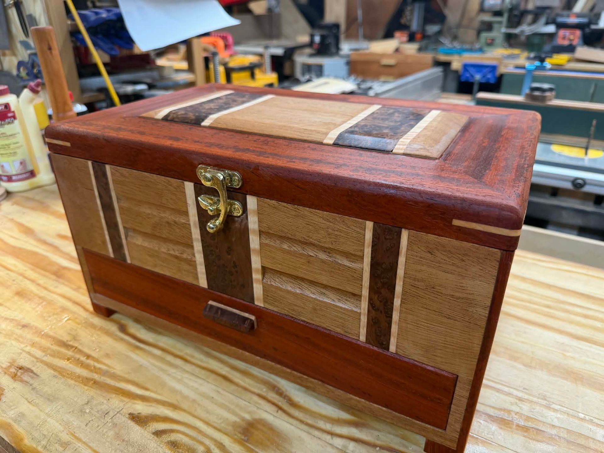 A handcrafted wooden box with a decorative, multi-toned inlaid lid and a brass latch, resting on a workbench.