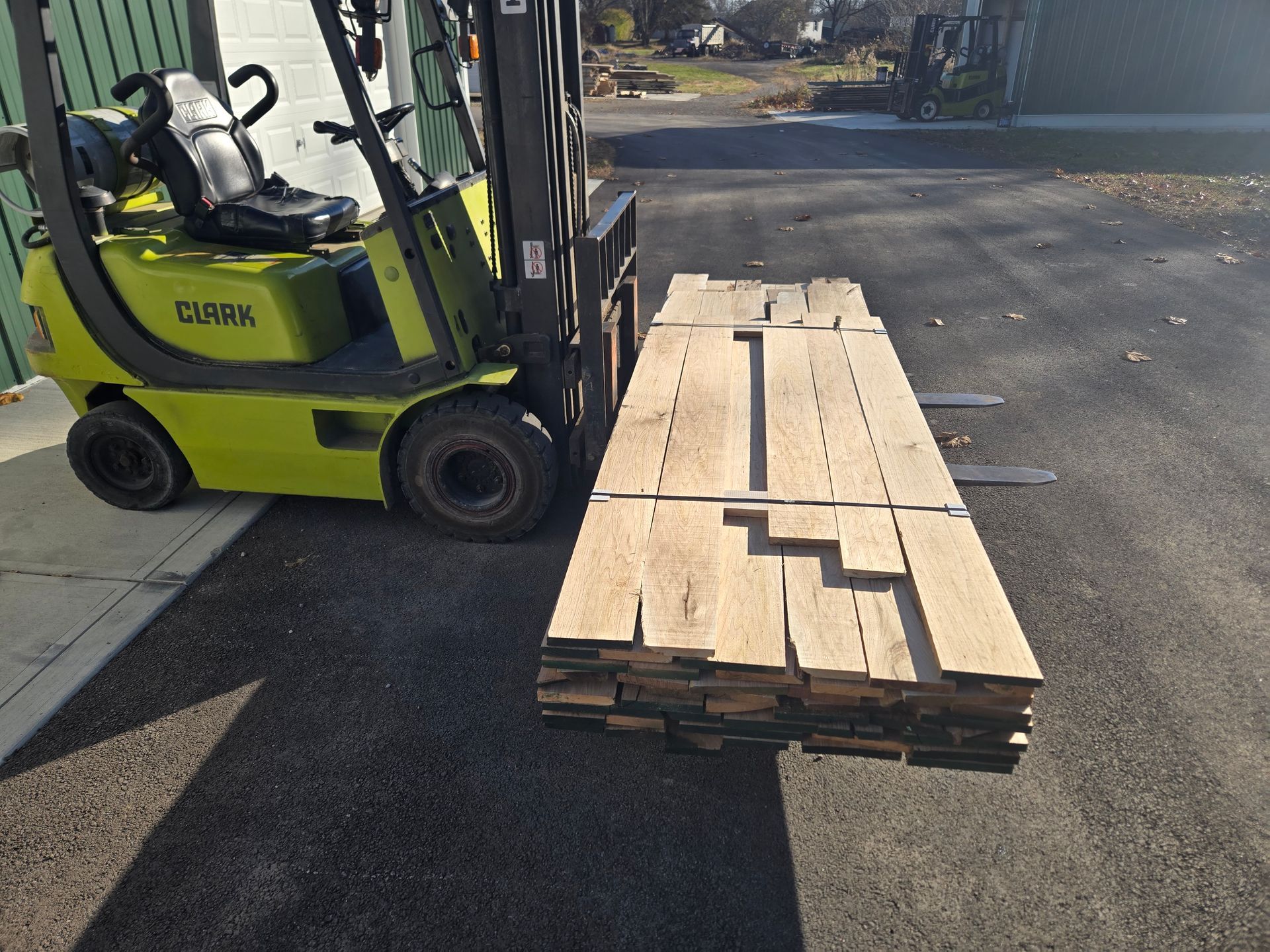 A yellow Clark forklift sits on pavement, its forks holding a large, neatly stacked bundle of light-colored wooden planks.
