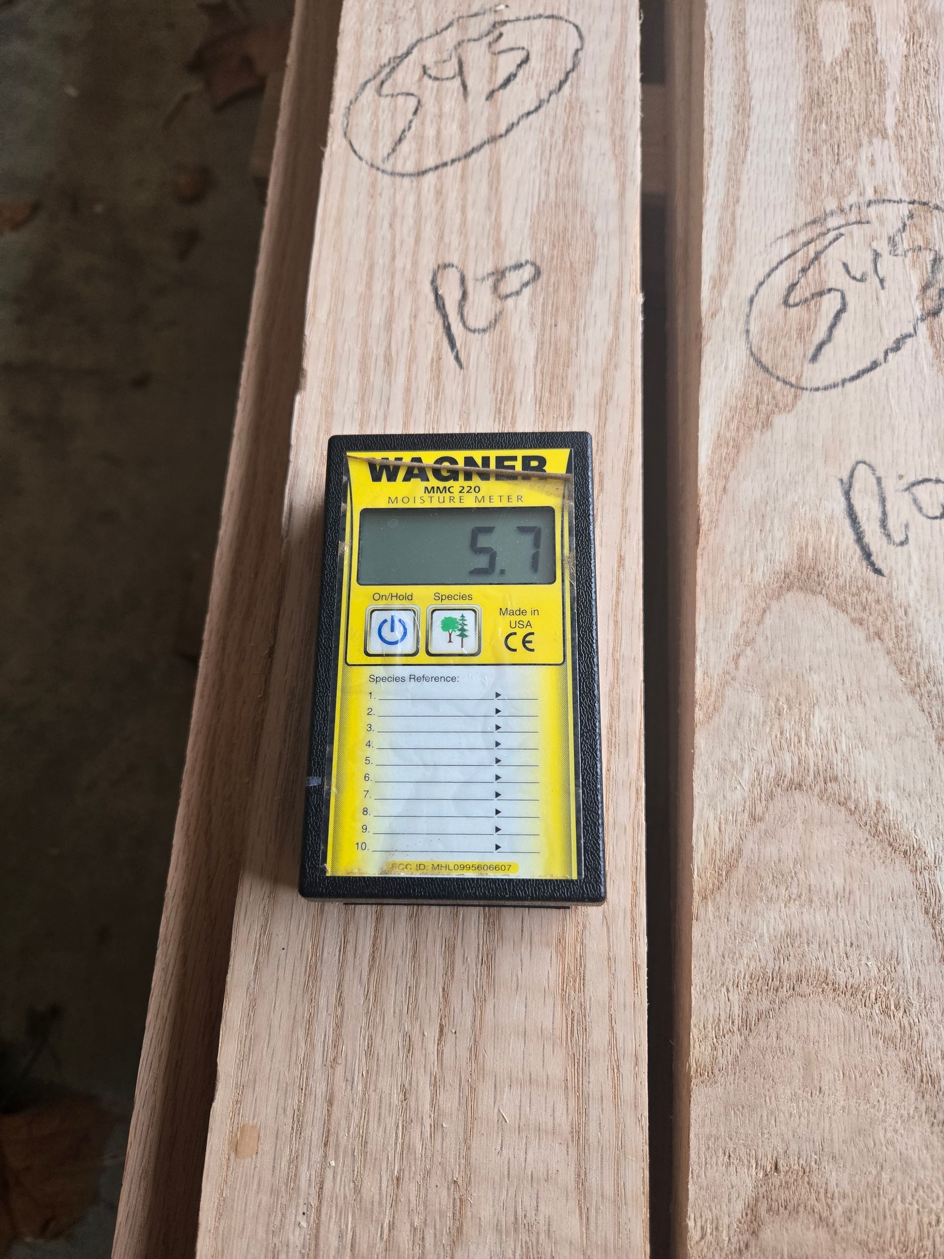 A yellow Wagner moisture meter showing a reading of 5.7 rests on a piece of light-colored, unfinished wood.