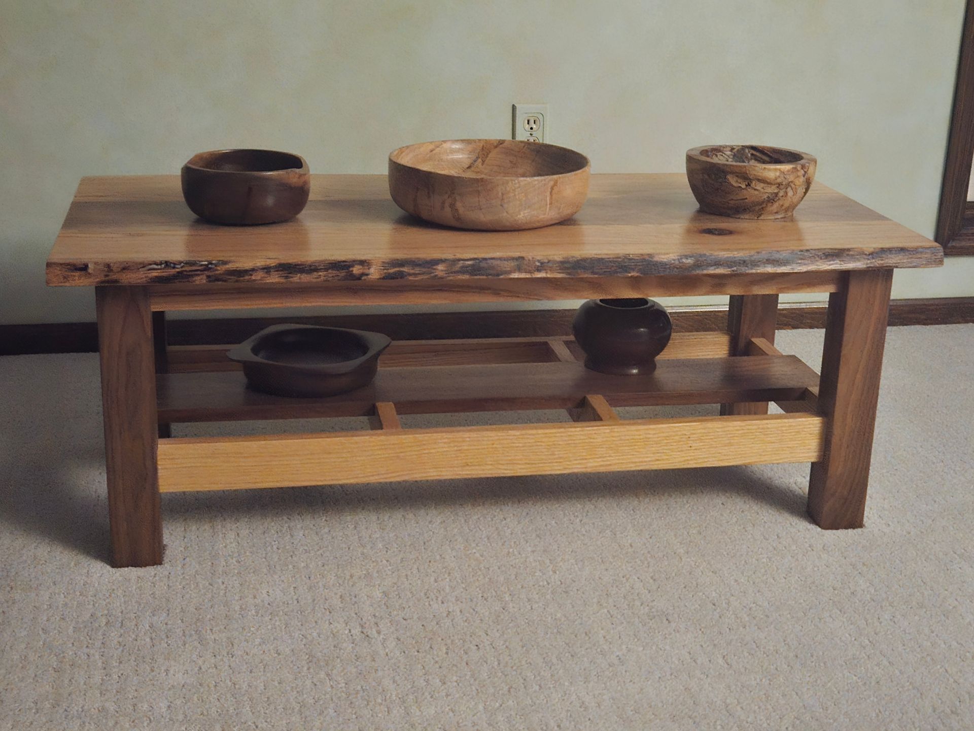 A live-edge wooden coffee table with three bowls on top and two objects on a lower shelf, set against a plain wall.