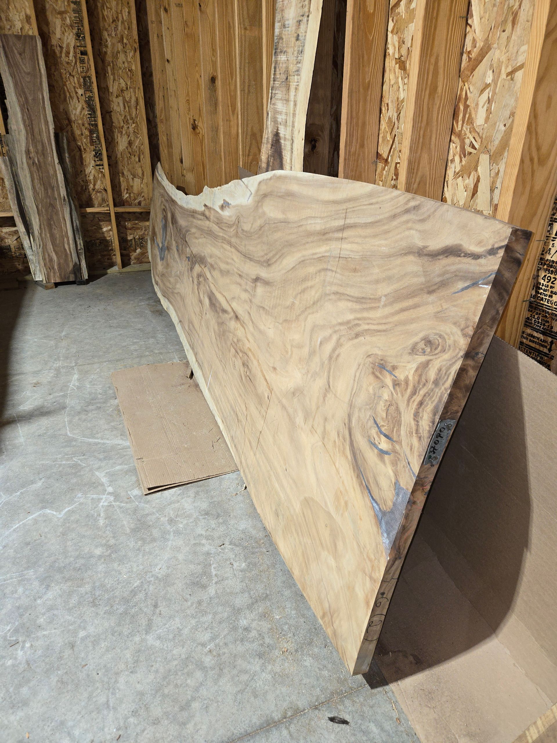 A large, natural-edge wood slab with swirling light and dark grain patterns leaning against a wall in a workshop.