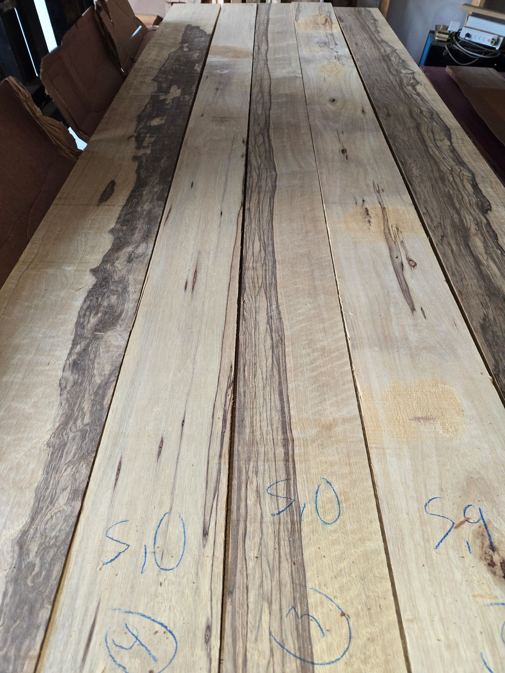 Several long, light-colored wooden planks with dark, textured live edges laid side-by-side on a workshop table.