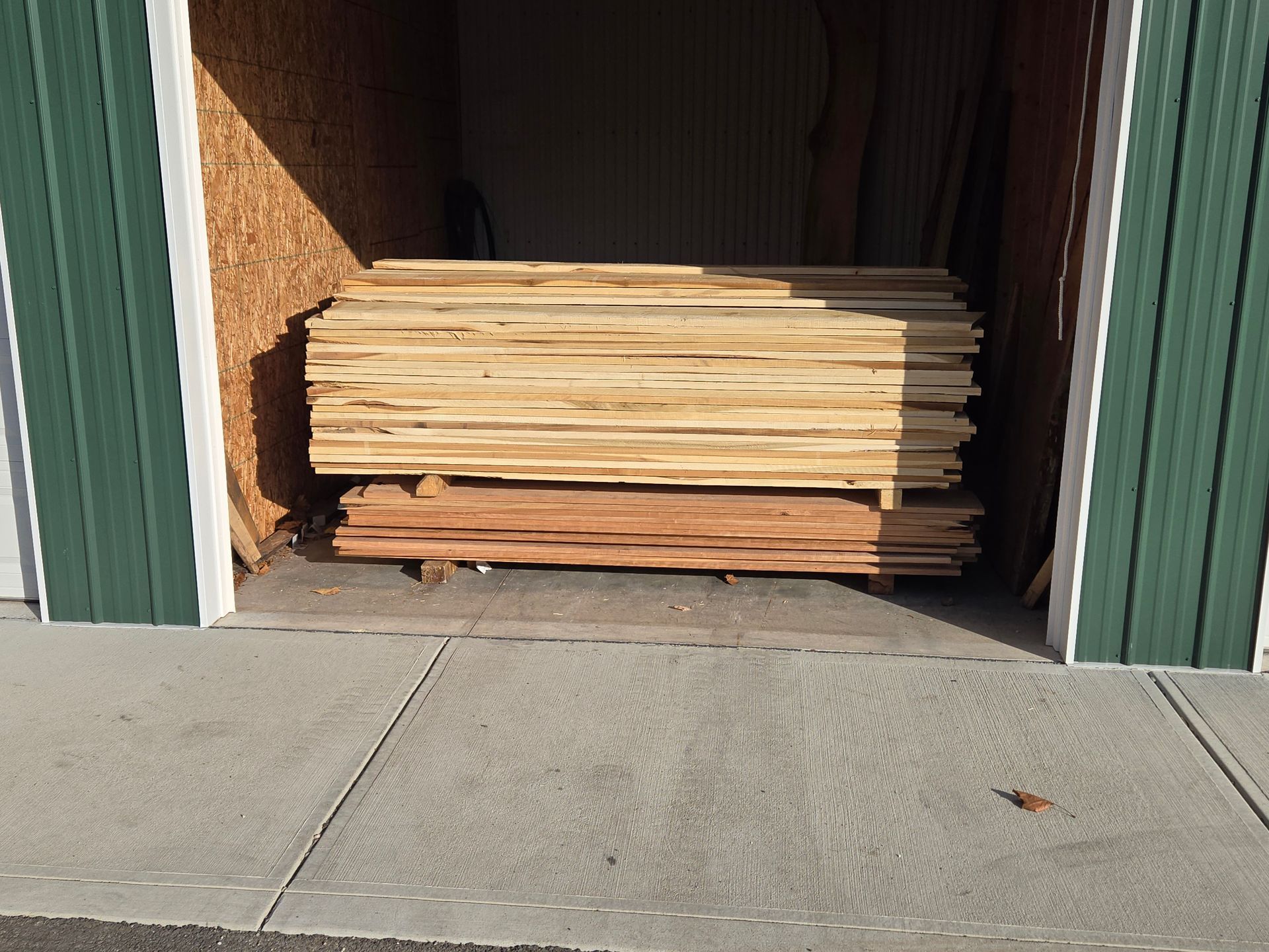 A stack of light and dark wooden boards stored inside a shed with green metal siding and a concrete floor.