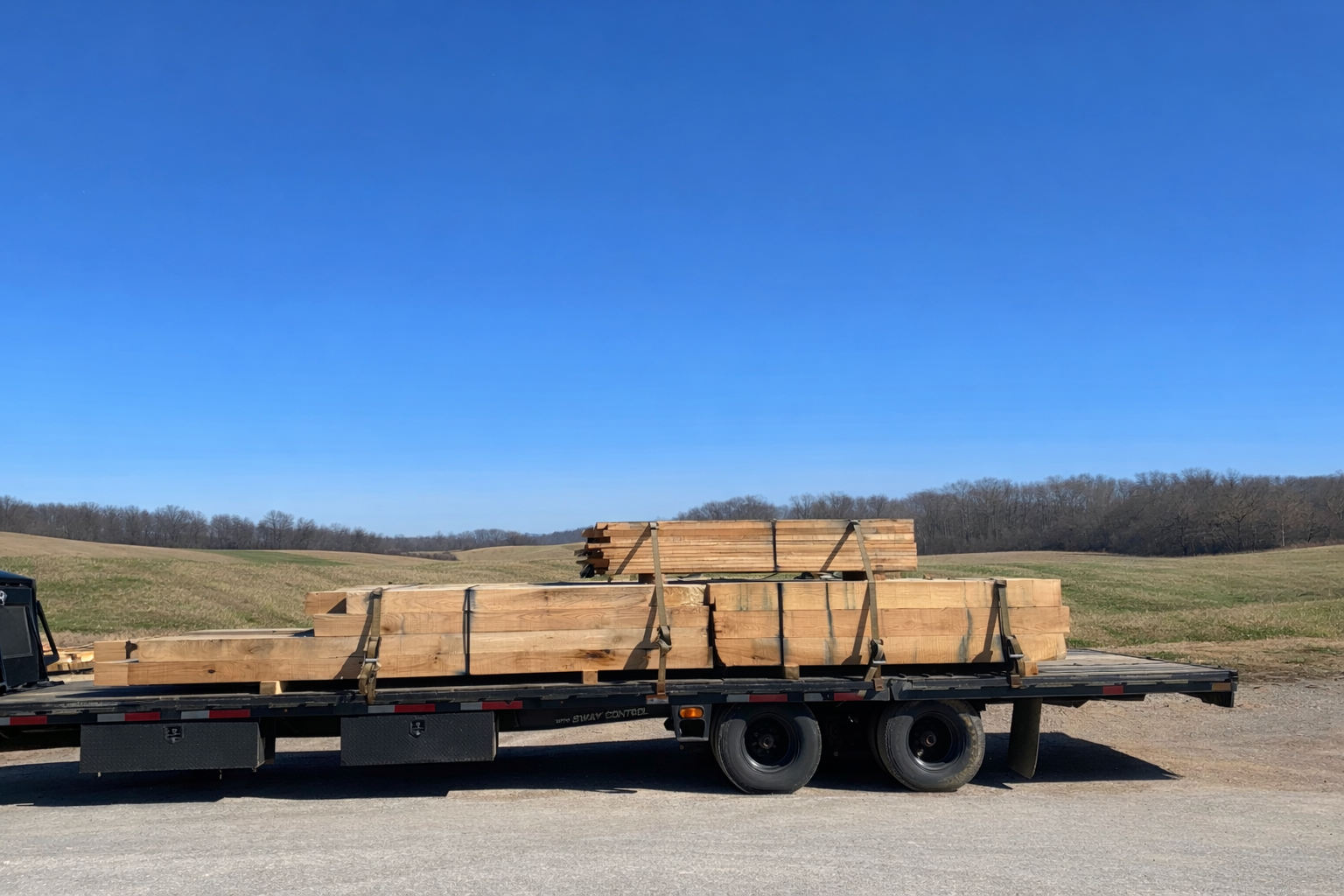 A flatbed trailer loaded with stacked, raw timber beams sits in a rural field under a clear blue sky.