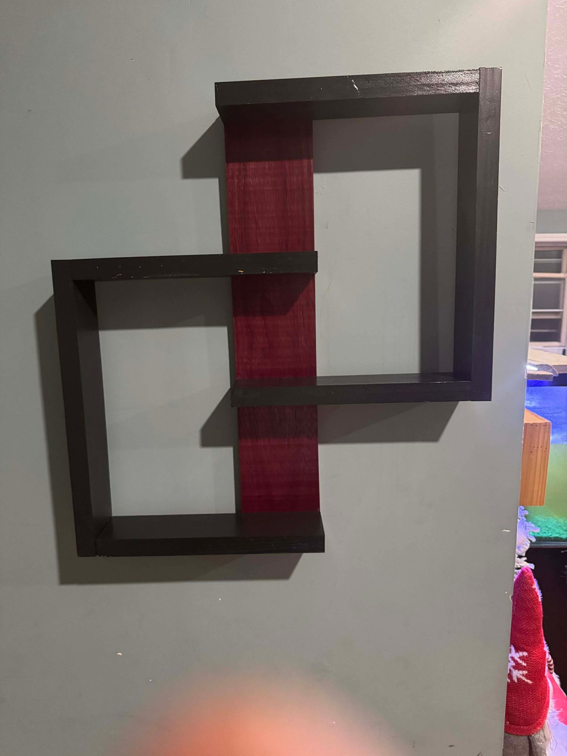 A wall-mounted geometric shelf with black frames and a vertical red center panel hanging on a light green wall.