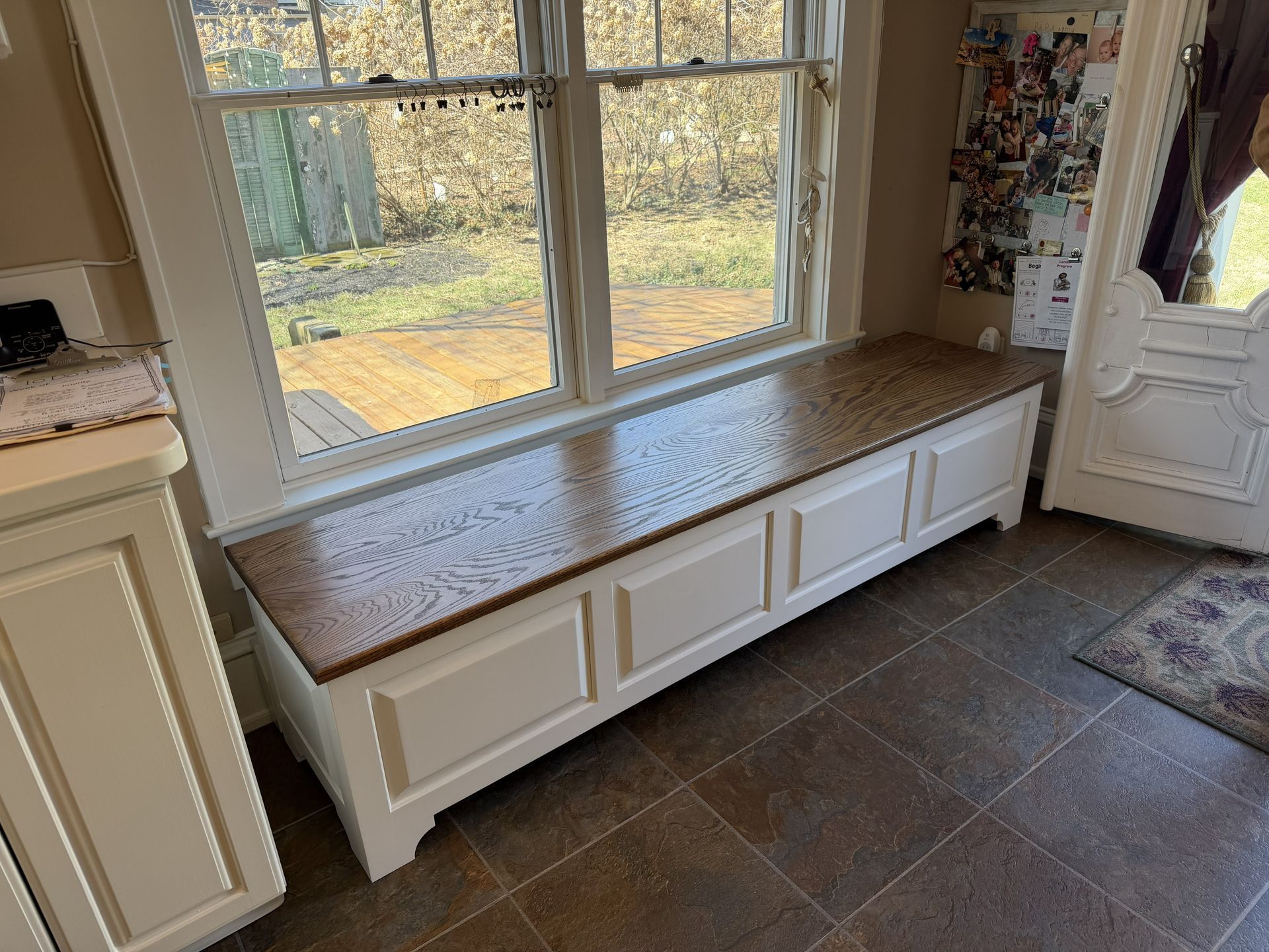 A white custom-built bench with a stained oak wood top sits below a large window in a tiled kitchen or entryway.