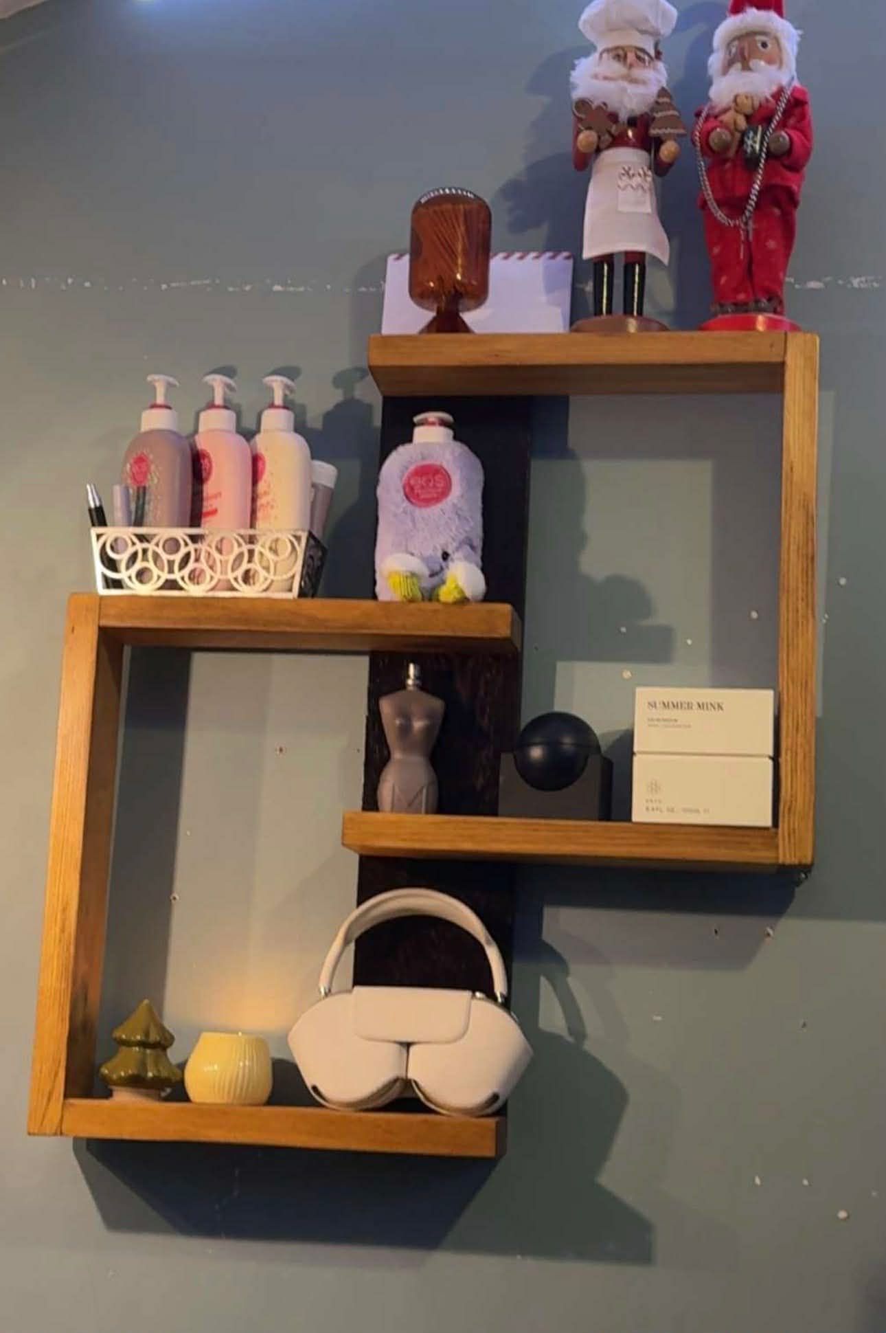 A wooden, multi-tiered wall shelf displaying skincare bottles, holiday figurines, a candle, and white headphones.