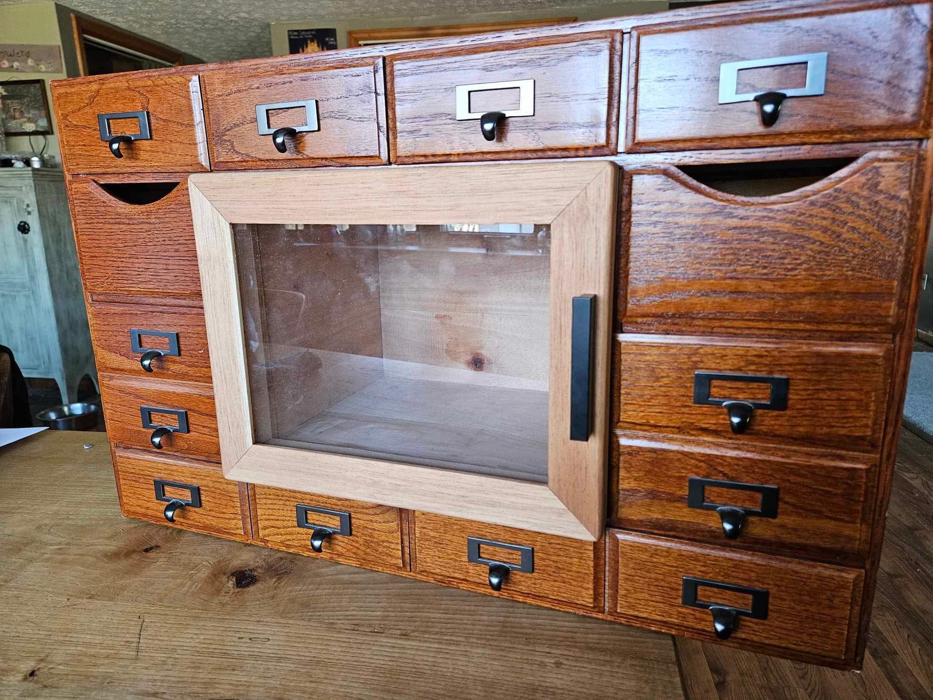A wooden multi-drawer storage unit with a central cabinet featuring a glass pane door and black metal hardware.