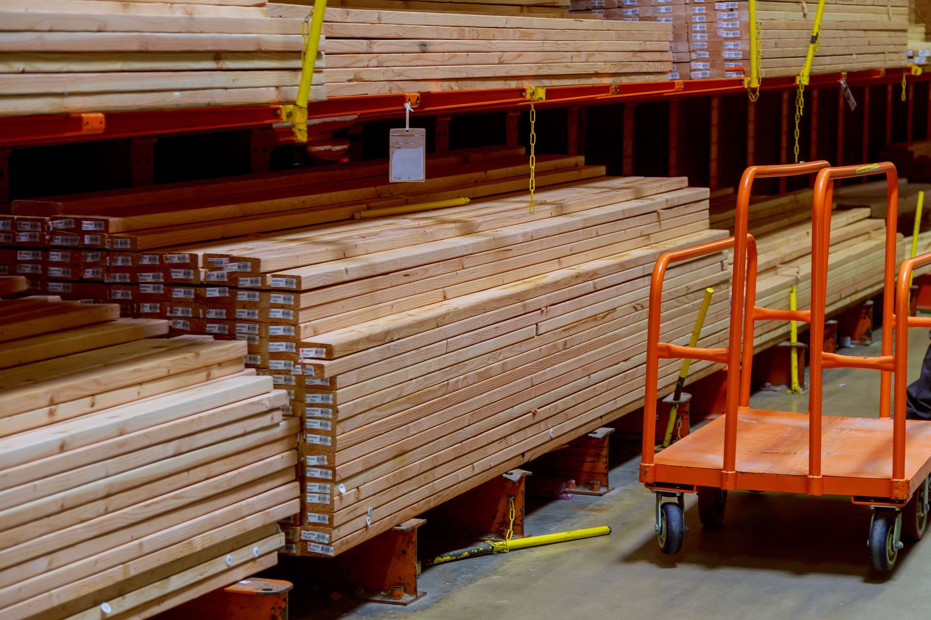 Stacked lumber piles in a hardware store aisle next to an empty orange metal flatbed cart.